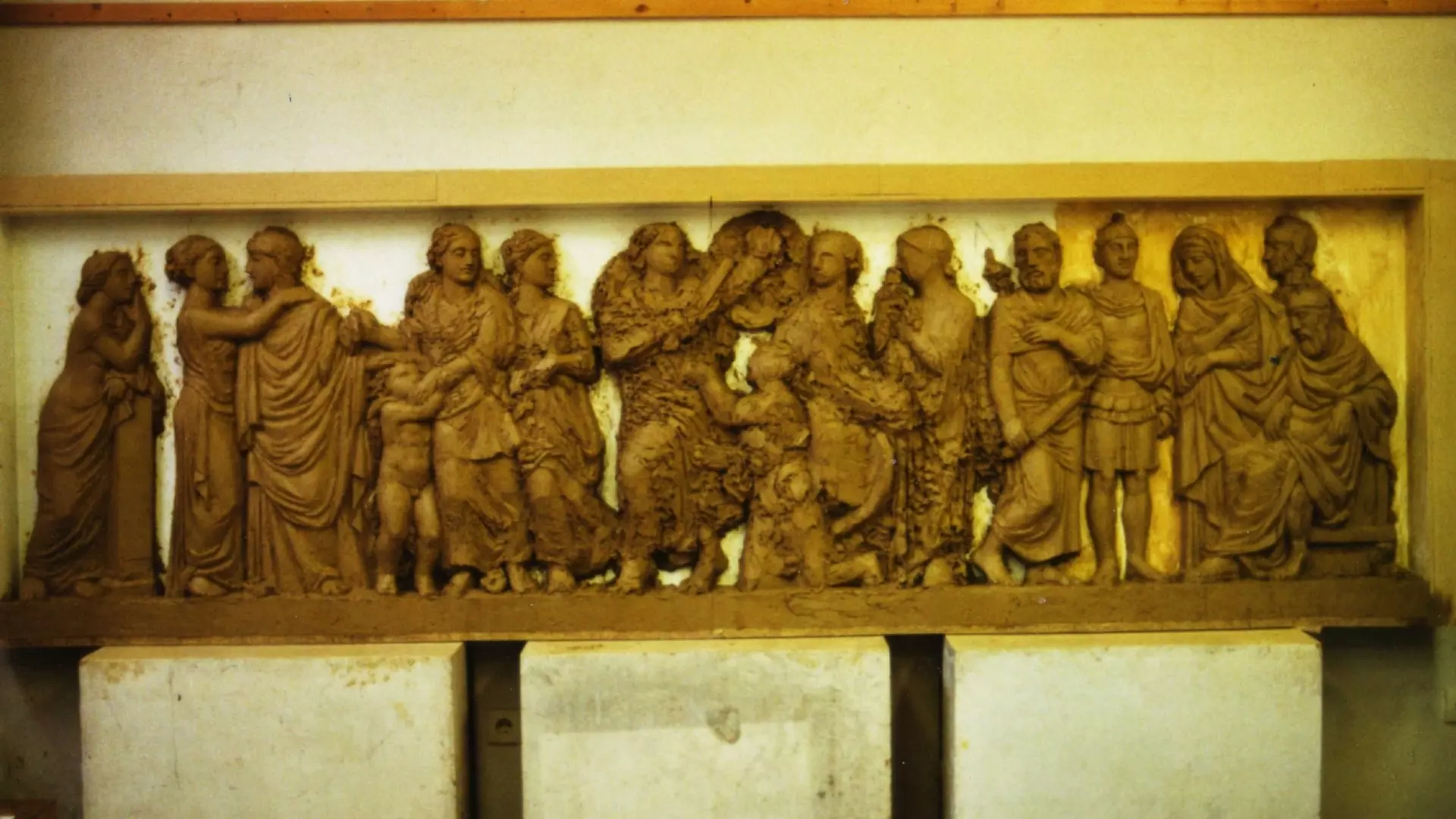 Langes, unfertiges Relief aus braunem Ton mit mehreren figürlichen Szenen, stehenden und sitzenden Personen in griechischen Gewändern, gelagert im Innenraum.