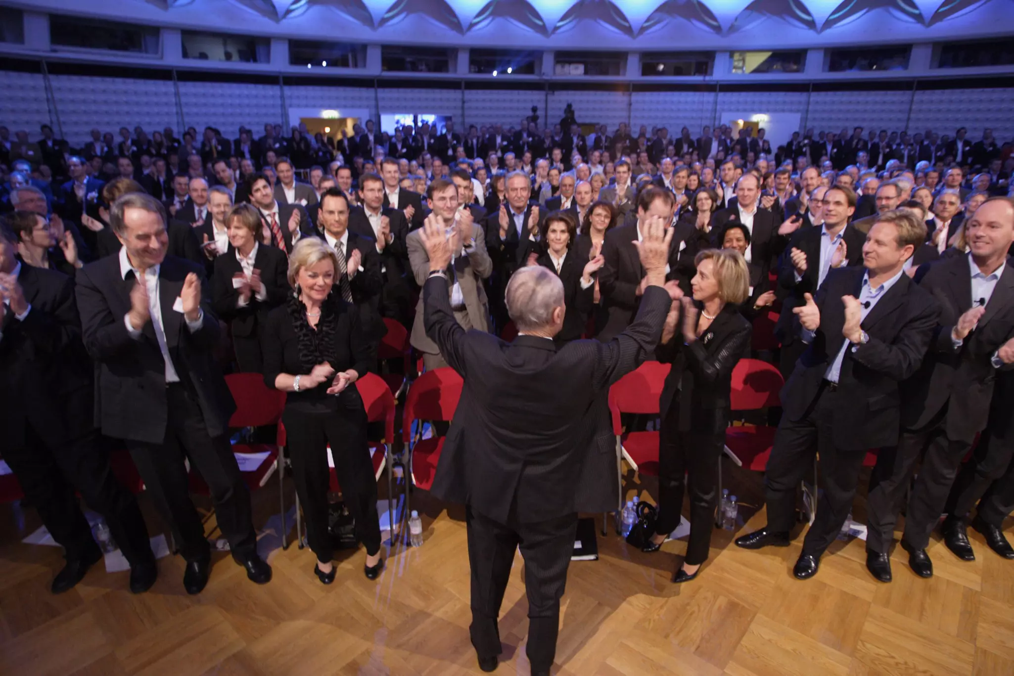 Reinhard Mohn faces a group of applauding people. He raises both hands.