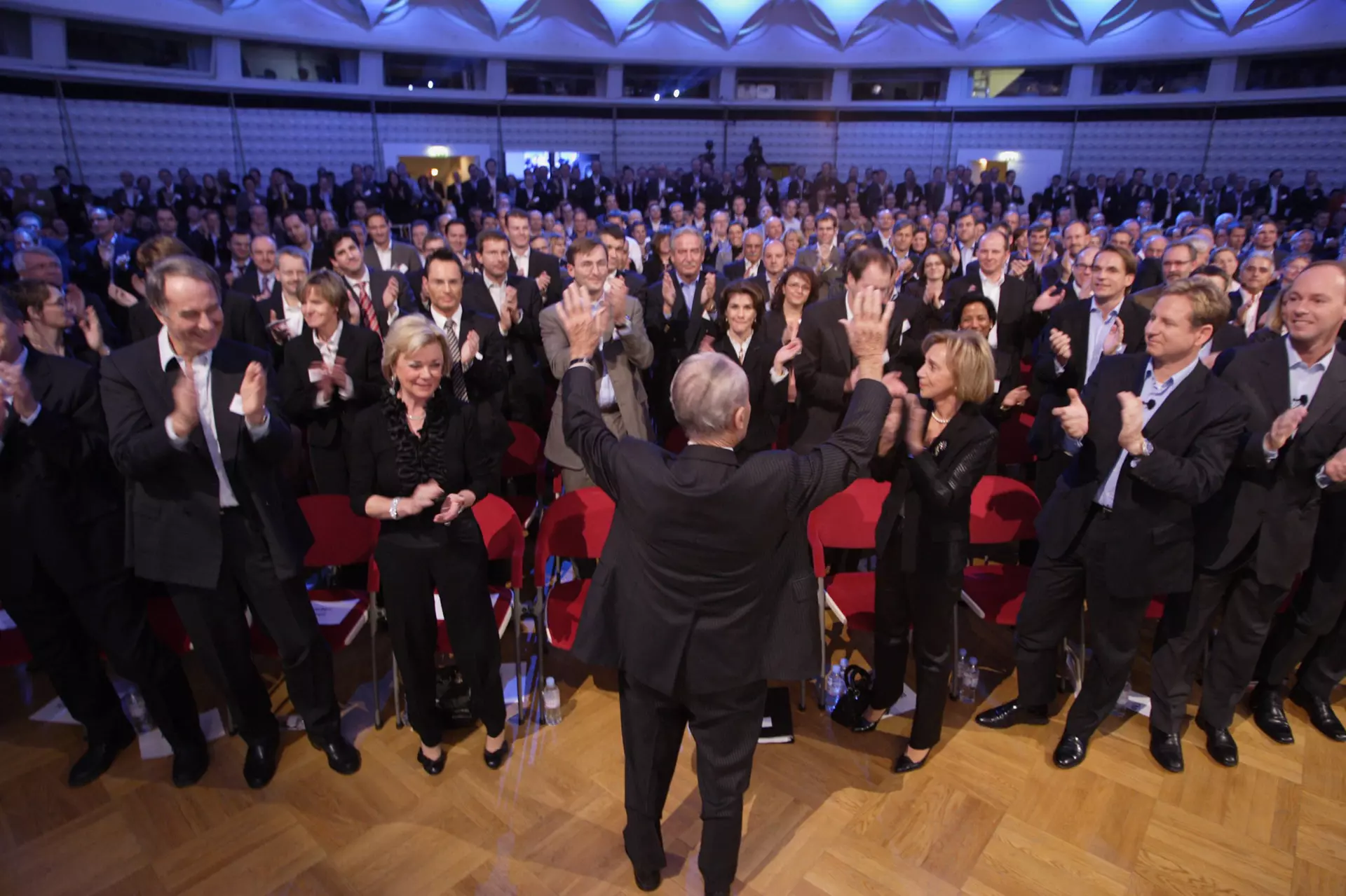 Reinhard Mohn faces a group of applauding people. He raises both hands.