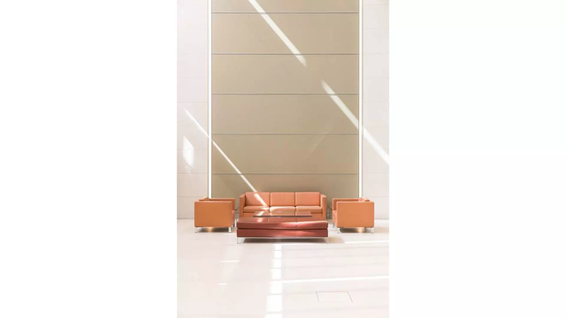 Sitzgruppe mit Sofa, Hocker und zwei Sesseln vor hoher Wand mit Metallpaneelen, Tageslicht fällt schräg durch Glasflächen.