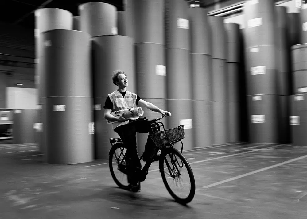 Person fährt Fahrrad mit Zeitungsrolle unterm Arm durch Industriehalle mit hohen Papierrollen im Hintergrund