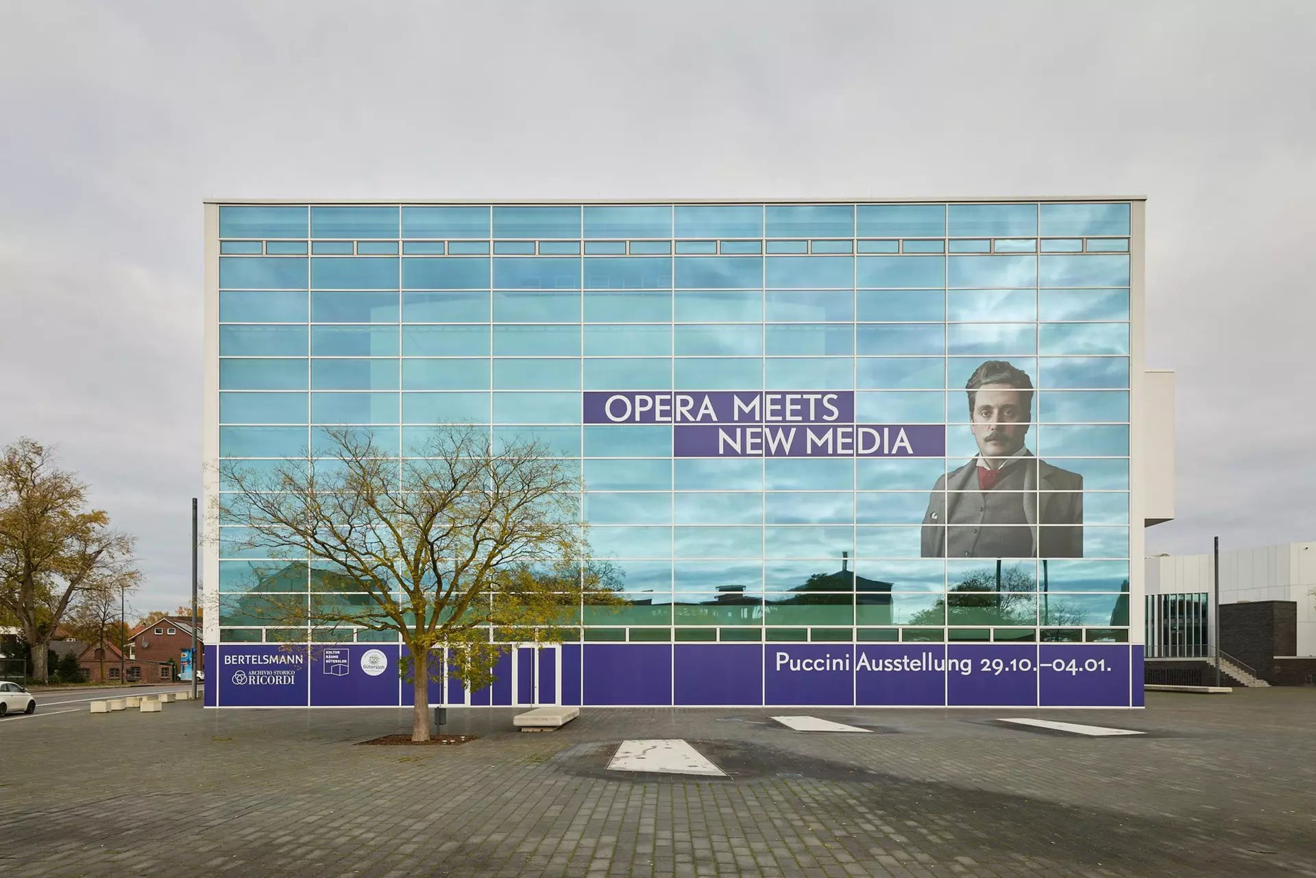 Exterior view of the Theater Gütersloh: The words “Opera Meets New Media” and a portrait of Giacomo Puccini are displayed prominently on the theater's exterior façade.