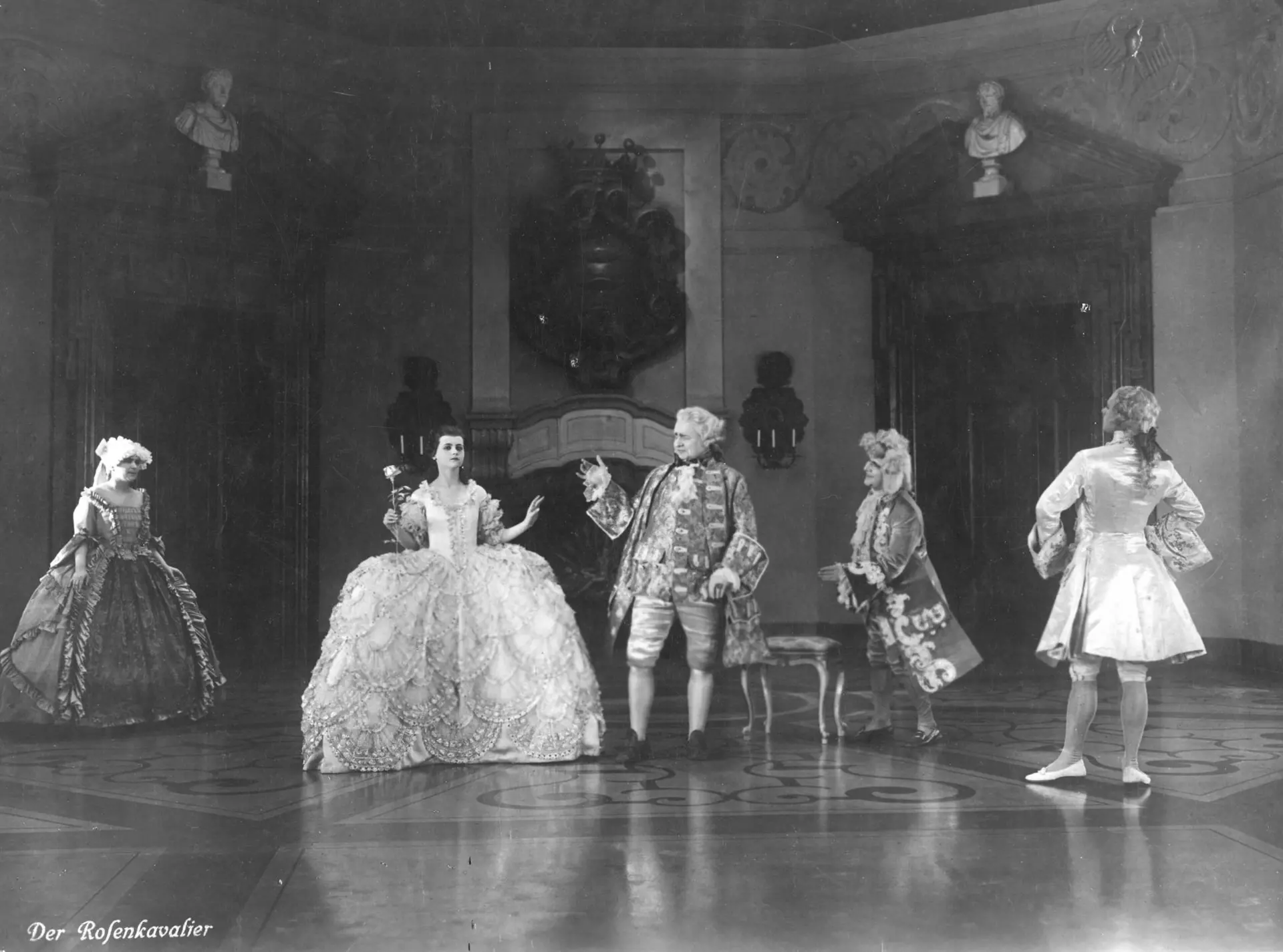 Ausschnitt aus dem Stummfilm "Der Rosenkavalier" in schwarz-weiß. Fünf Personen stehen in einer Filmkulisse, einem großen Saal eines Schlosses. Die Personen tragen Kleidung und Perücken aus dem Zeitalter des Rokokos. Eine in einem ausladenden Kleid gewandete Person hält eine Rose in der Hand. Unten links steht in weißer Schrift geschrieben: "Der Rosenkavalier".