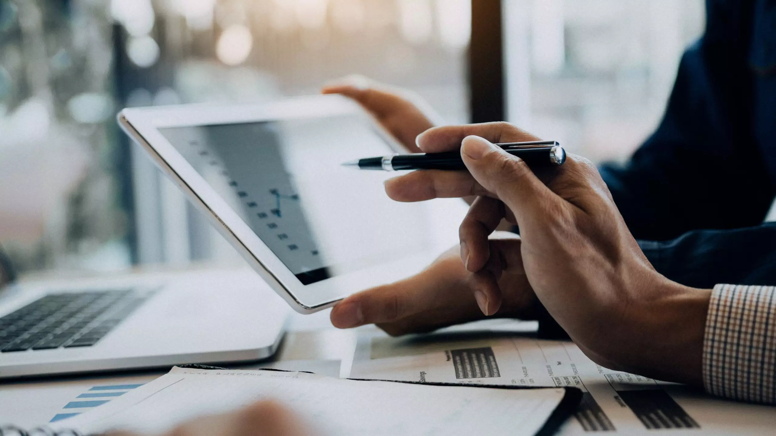 Hand pointing at a tablet with a financial chart, with a laptop and documents beside it.