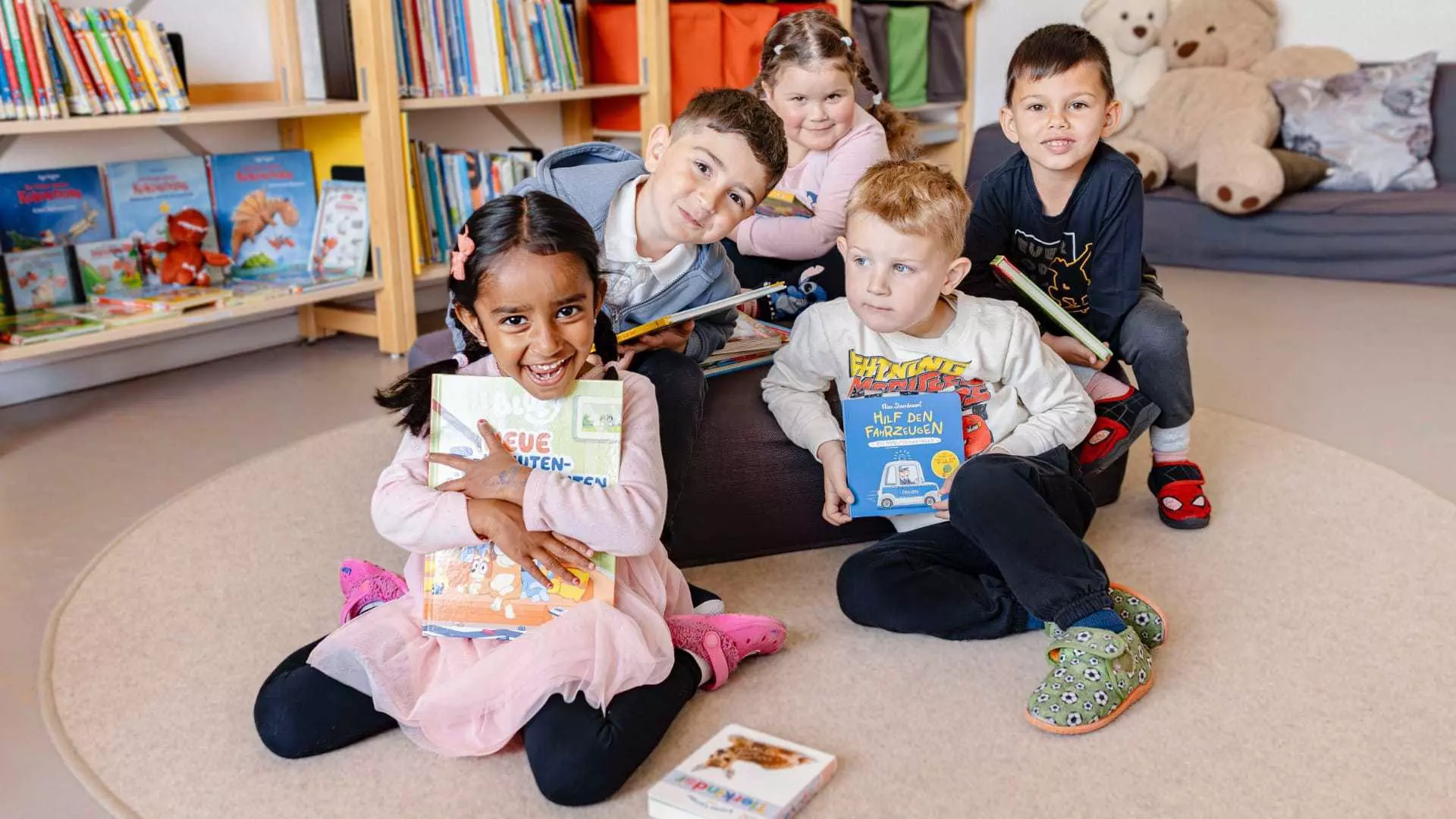 Fünf Kinder sitzen im Gruppenraum, halten Bücher und schauen in die Kamera, umgeben von Regalen und Spielzeug.