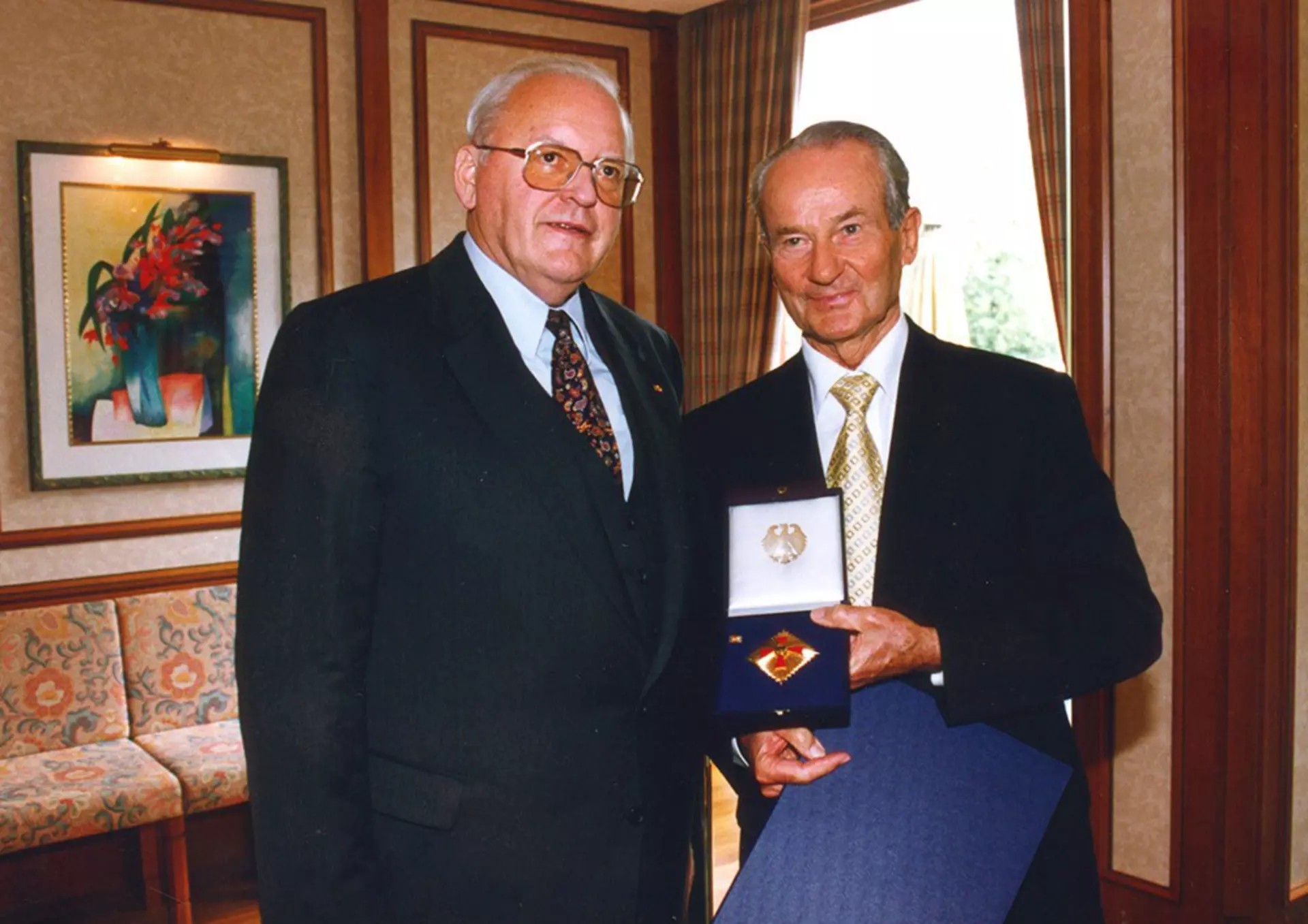 Reinhard Mohn stands next to Roman Herzog and holds a box containing the Federal Cross of Merit and a blue folder.