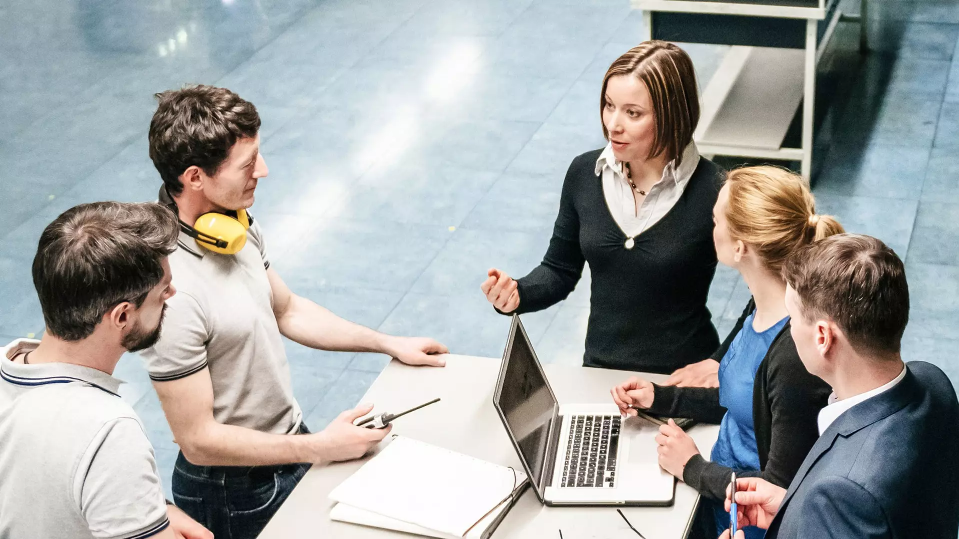 Five people are standing around a table discussing a project; a woman is explaining something while the others listen, take notes, and look at an open laptop.