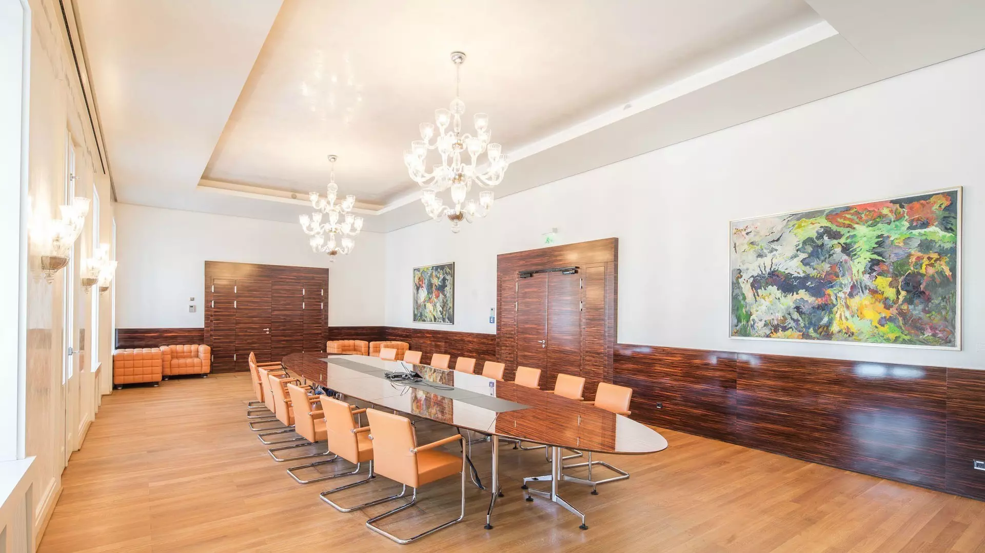 Eleganter Konferenzraum mit langem ovalen Tisch, lederbezogenen Stühlen, Kronleuchtern, Holzwänden und großformatigen Gemälden.