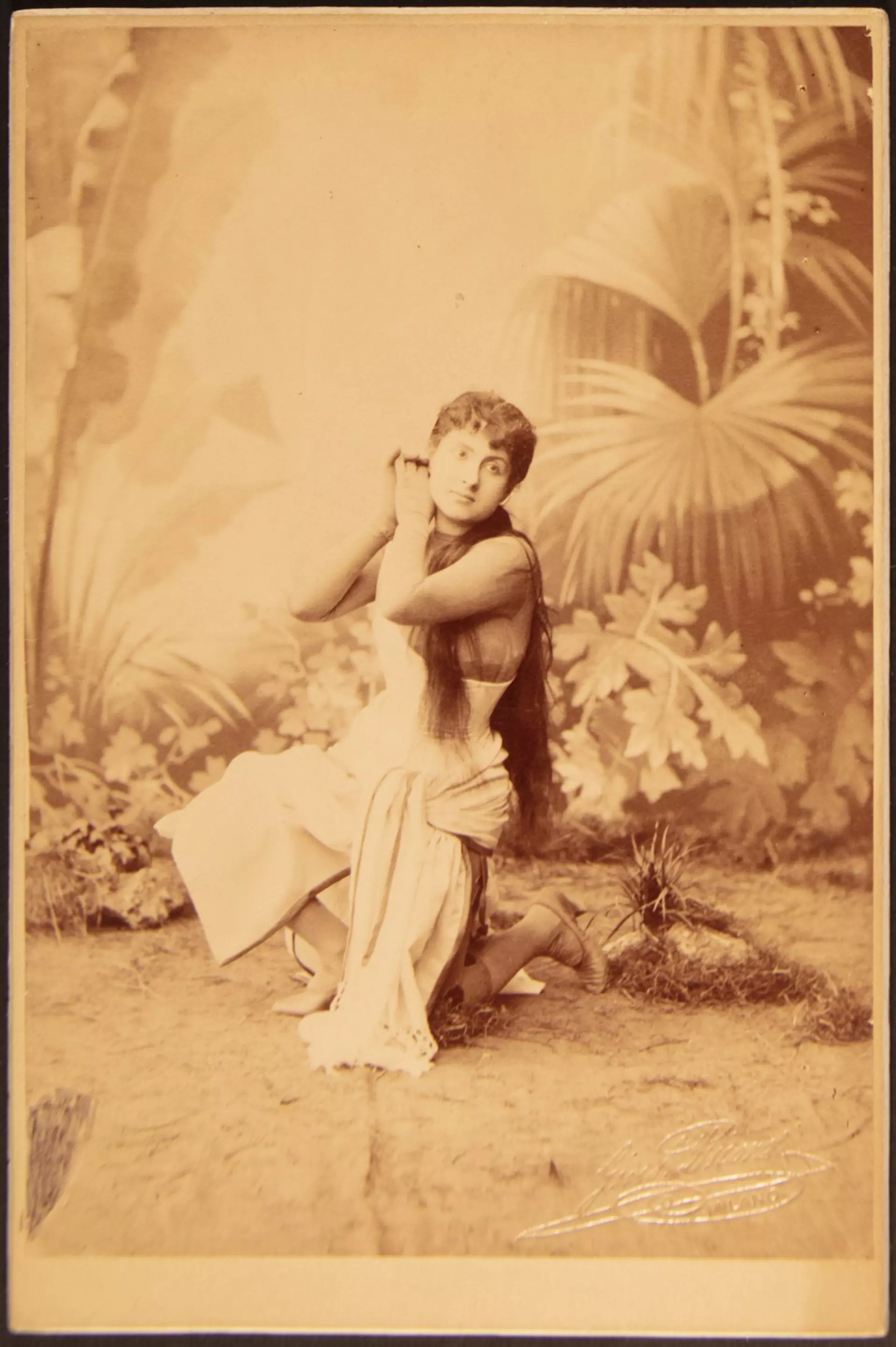 Historisches Porträt einer knienden Tänzerin in langem Kleid vor gemaltem Pflanzenhintergrund.