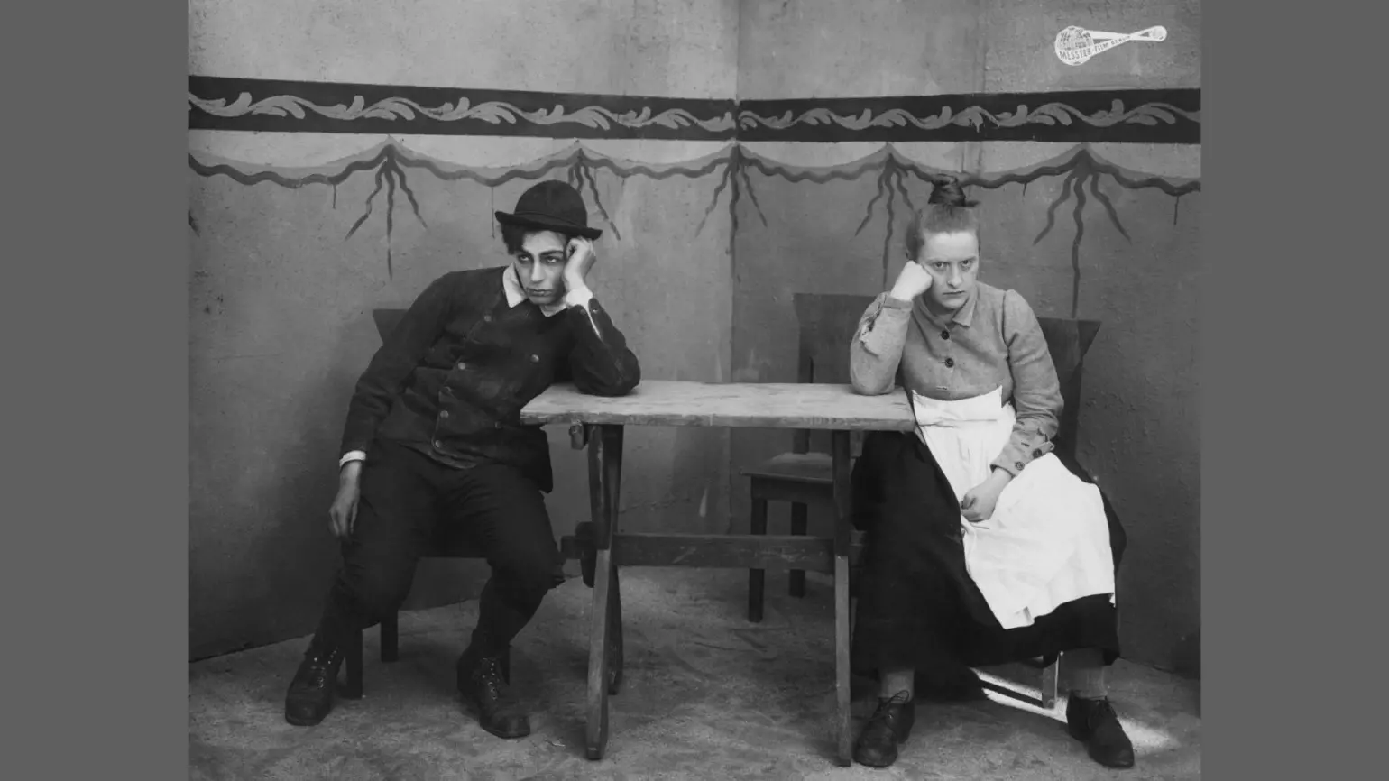 Schwarzweiß-Filmstill mit einem Mann und einer Frau an einem einfachen Holztisch. Beide sitzen, in unterschiedliche Richtungen guckend, den Kopf in die Hand gestützt. Der Raum ist karg, der Hintergrund zeigt eine bemalte Wand mit ornamentalen Motiven.