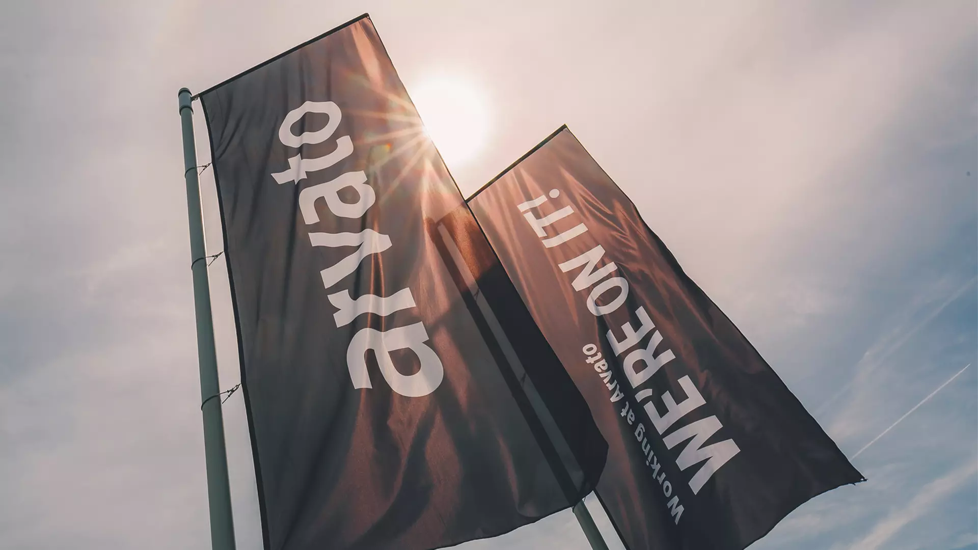 Two flags are fluttering in the wind under the sun. They bear the inscriptions: Arvato and We're on it.