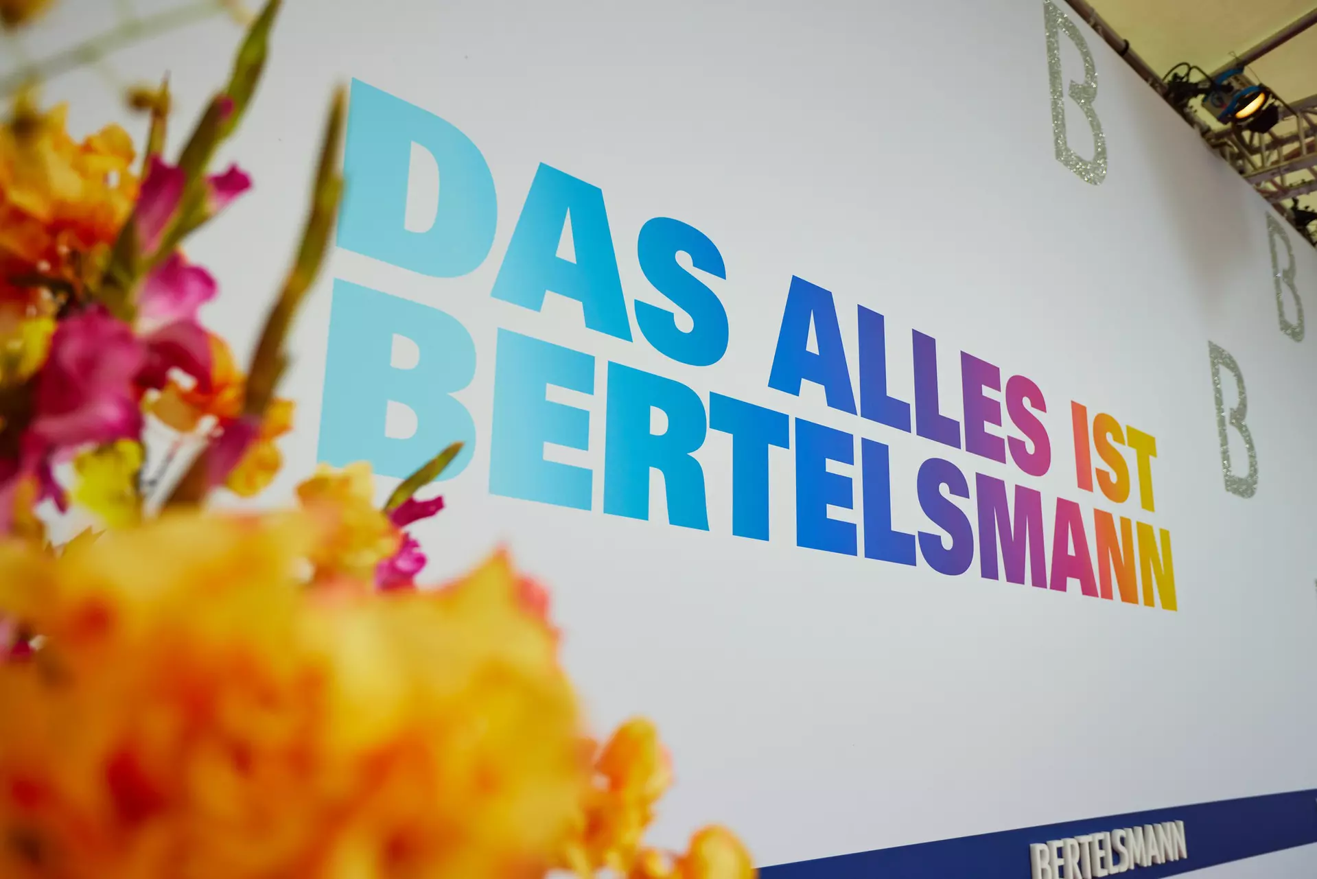 Fotowand am roten Teppich mit bunter Inschrift "Das alles ist Bertelsmann", drei silberglitzernden Bs und dem Bertelsmann Logo. Im Vordergrund sieht man unscharf einen Teil eines gelb-pinken Blumenbouquets.