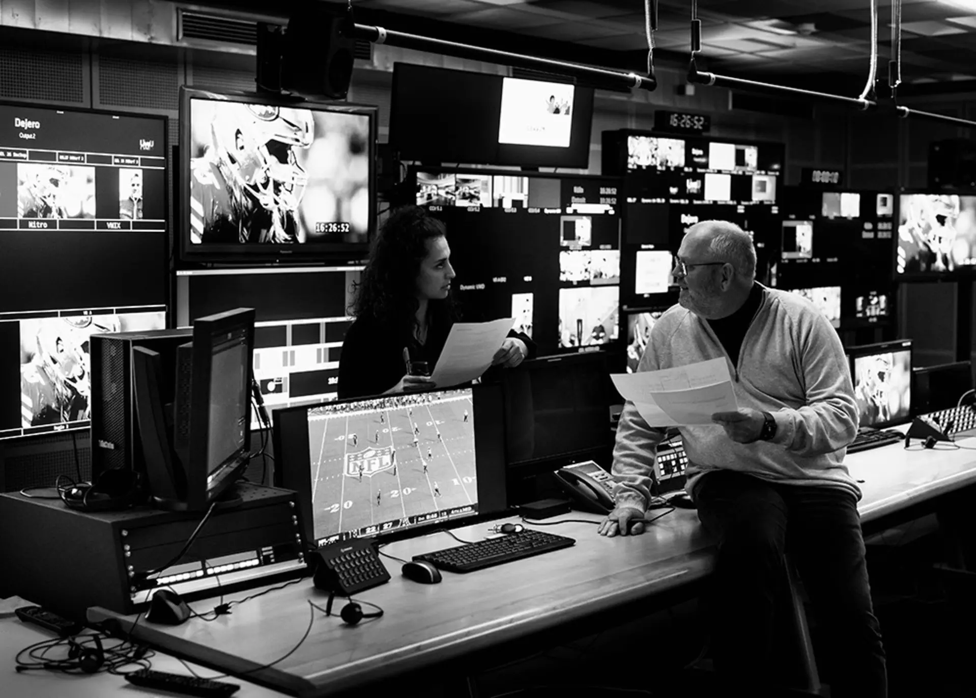 Zwei Personen im Videokontrollraum unterhalten sich, im Hintergrund viele Bildschirme mit NFL-Szenen