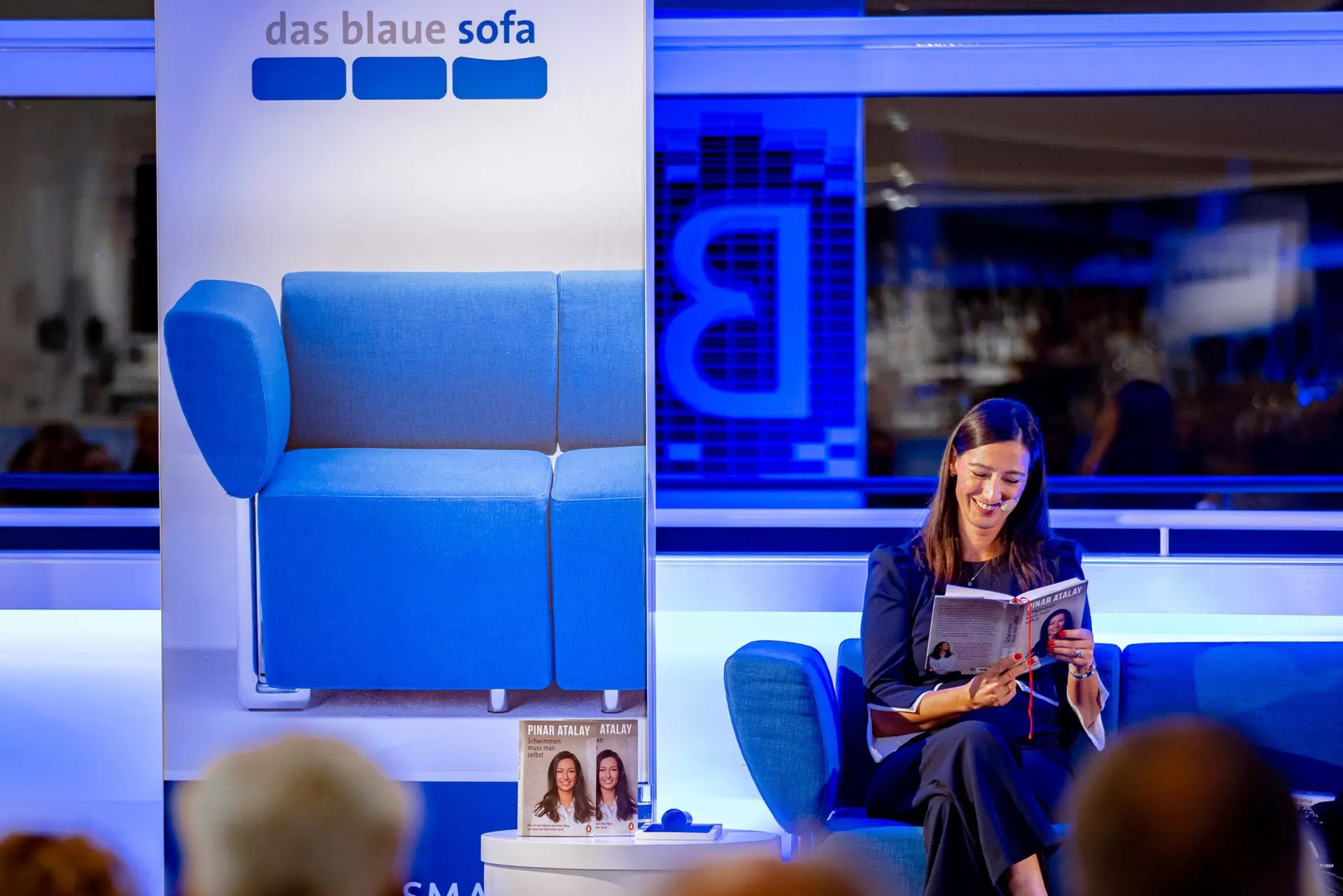 Die Autorin sitzt auf dem blauen Möbelstück und liest aus ihrem aufgeschlagenen Buch. Im Vordergrund sieht man unscharf die Hinterköpfe von Personen aus dem Publikum.