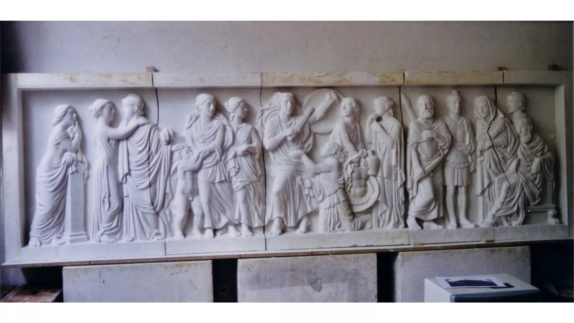 Langes Relief aus weißem Stein mit mehreren figürlichen Szenen, stehenden und sitzenden Personen in griechischen Gewändern, gelagert im Innenraum.