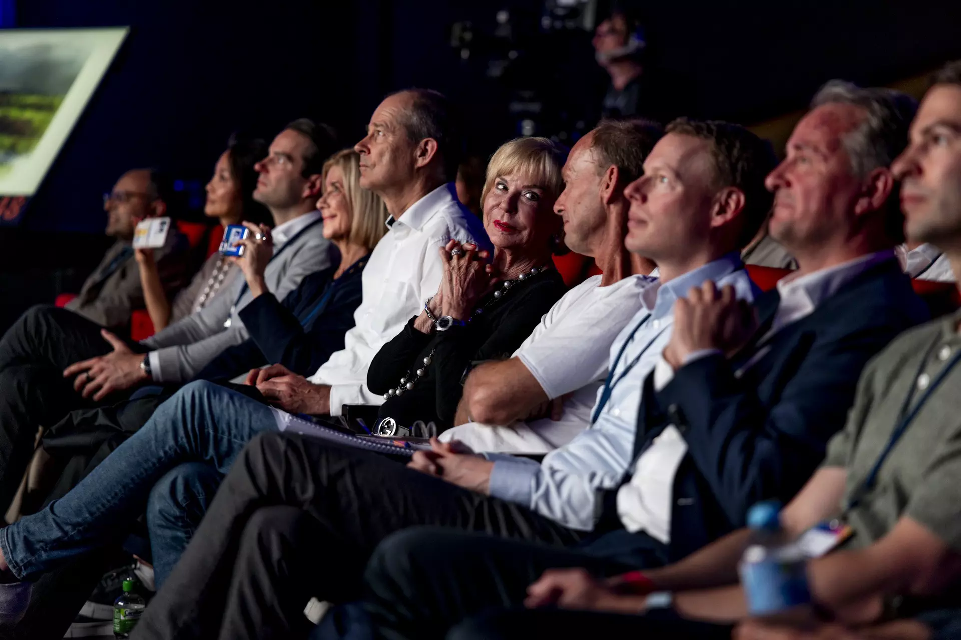 Mehrere Personen sitzen in vorderer Reihe eines Auditoriums und verfolgen aufmerksam eine Bühnenpräsentation.