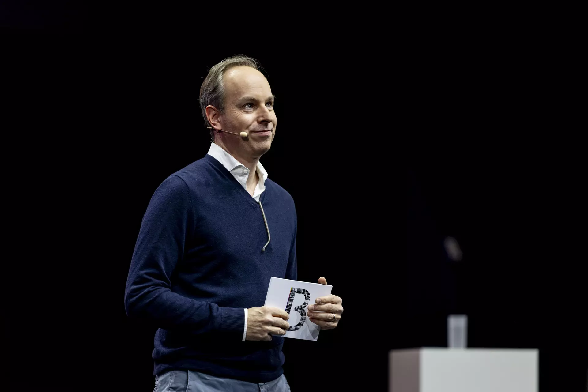 Person mit Headset hält Karte in der Hand und spricht auf Bühne vor dunklem Hintergrund.