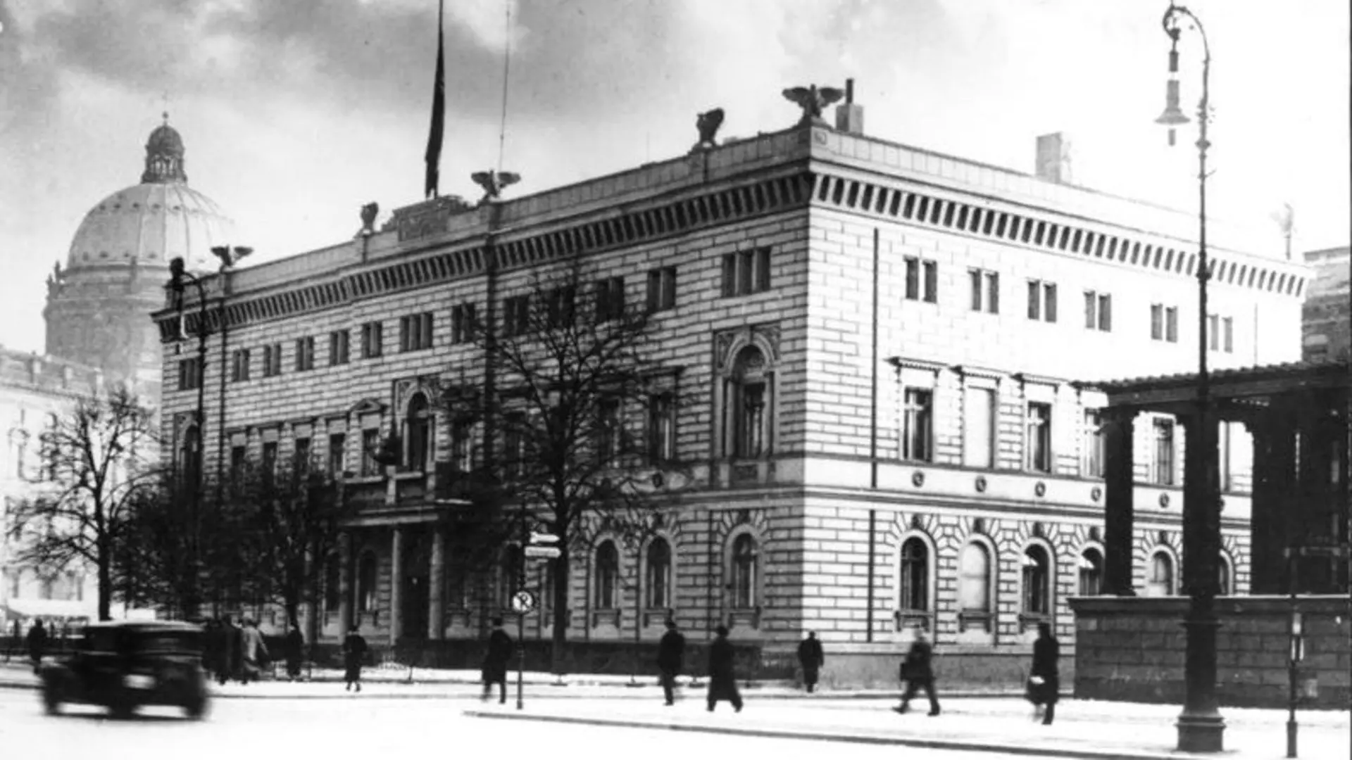 Historische Fotografie von 1937 mit großem klassizistischem Eckgebäude, Bäume davor Passanten und alten Automobilen  auf breiter Straße.
