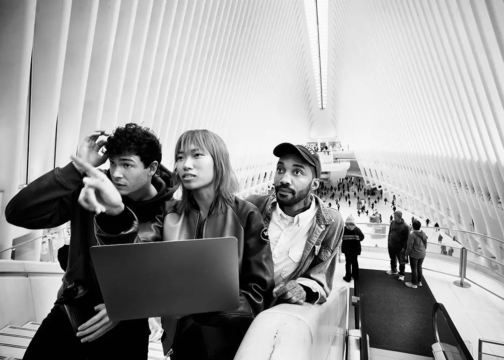 Drei Personen mit Laptop auf Rolltreppe in heller Halle mit symmetrischer Architektur.