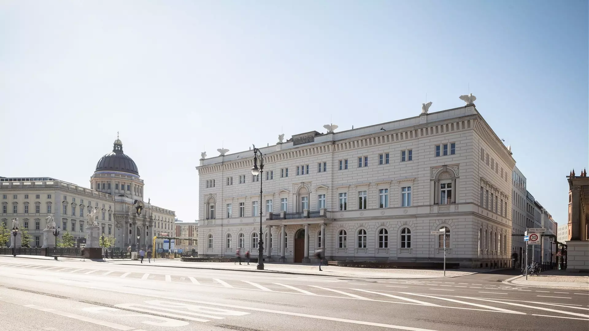 Breite Straße, helles klassizistisches Eckgebäude mit großem Balkon im 1. Stock und Adlerfiguren auf dem Dach, im Hintergrund Kuppelbau, wenige Passanten.