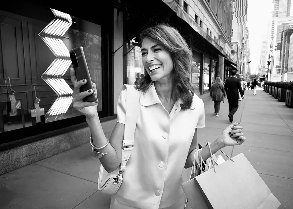 Person in Einkaufsstraße vor Schaufenstern schaut lächelnd ins Smartphone und trägt mehrere Einkaufstüten