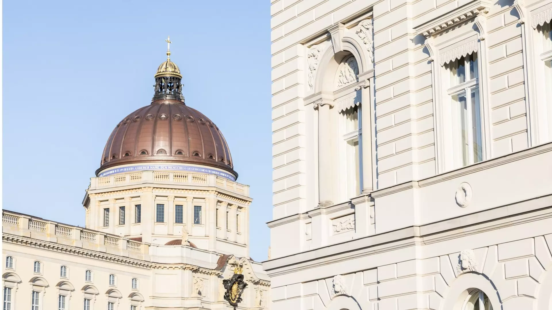 Blick auf kupfernen Kuppelbau mit Kreuz und vergoldeten Reliefs, rechts helle klassizistische Fassade mit Fenstern und Reliefs.