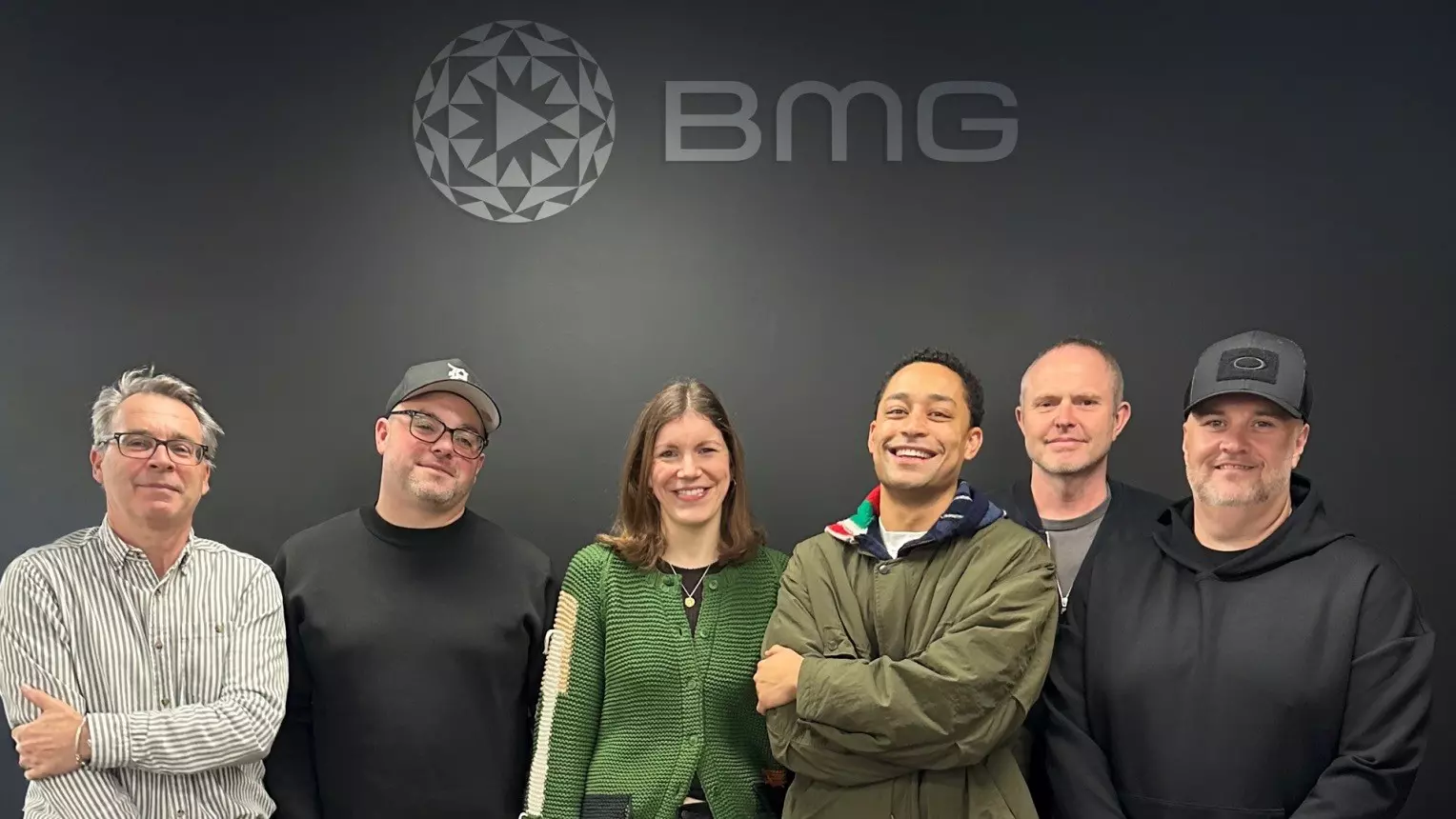 Five men and one woman stand in front of a gray wall with a light gray BMG logo. They are all looking at the camera, some of them smiling. They are Alistair Norbury, Chris Wareing, Clemmie Woodhouse, artist Loyle Carner, Hugo Turquet, and Ben Kaelin.