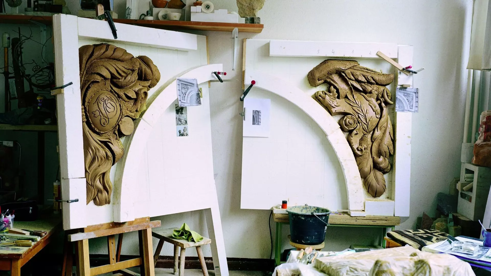 Werkstatt mit zwei bogenförmigen Schmuckelementen aus ungebranntem Ton, in Schablonen fixiert, Werkzeuge im Raum.