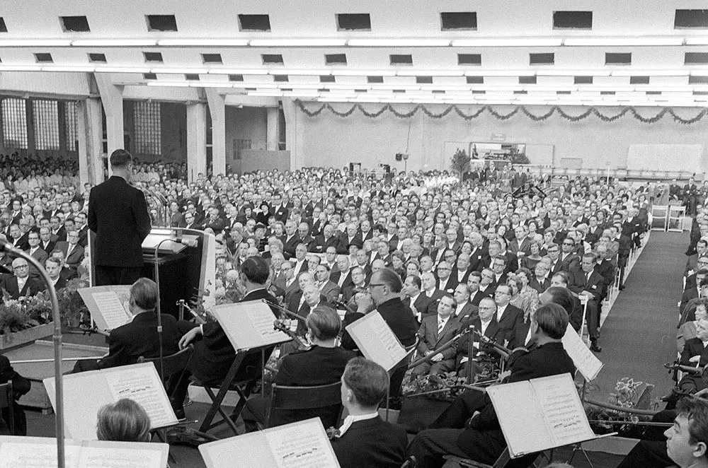 Reinhard Mohn stands at a lectern facing a large number of guests seated in a decorated ballroom. Behind him sit members of an orchestra with instruments and sheets of music.