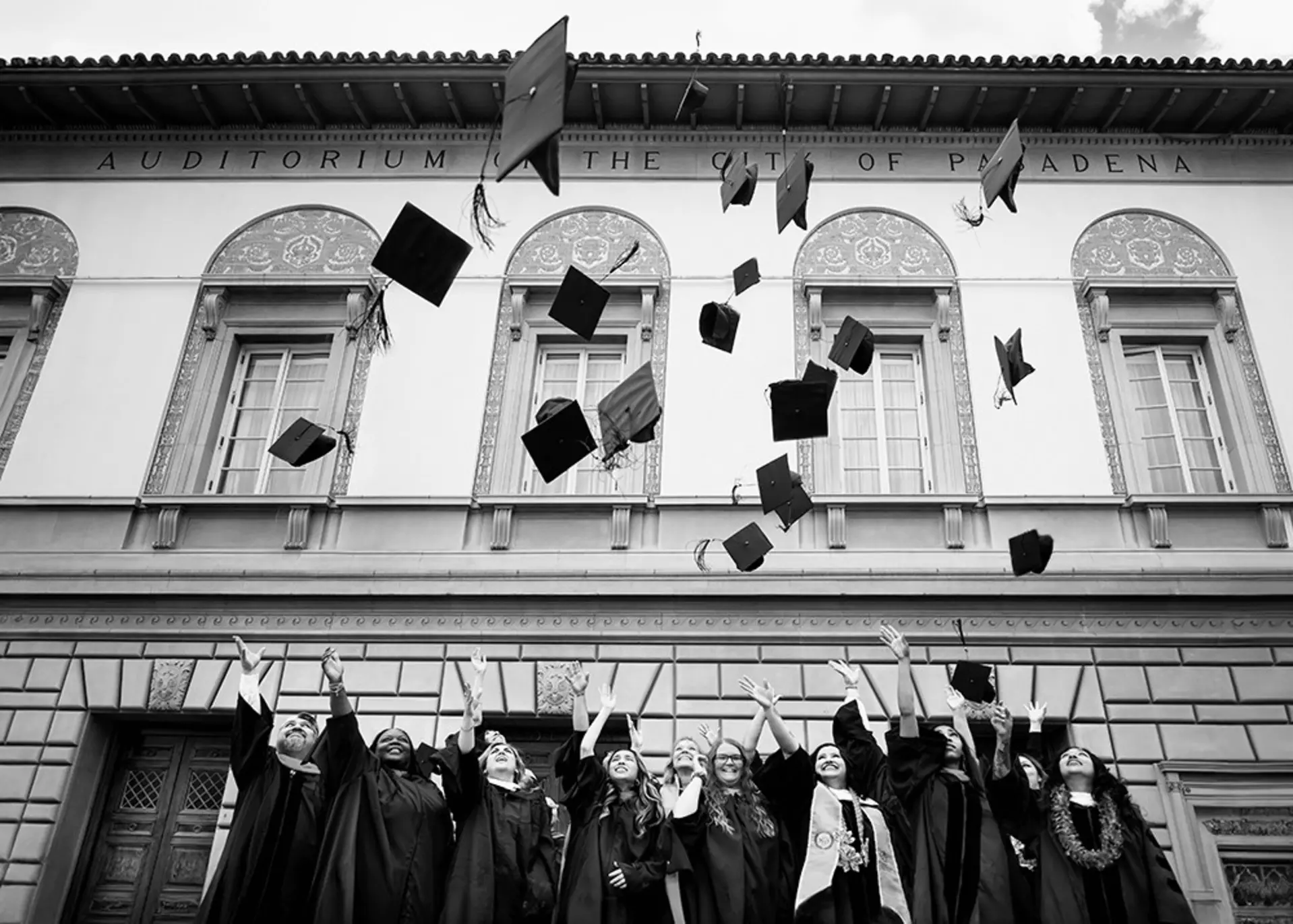 Absolventinnen und Absolventen werfen Absolventenhüte vor historischer Fassade mit Schriftzug „AUDITORIUM OF THE CITY OF PASADENA“.