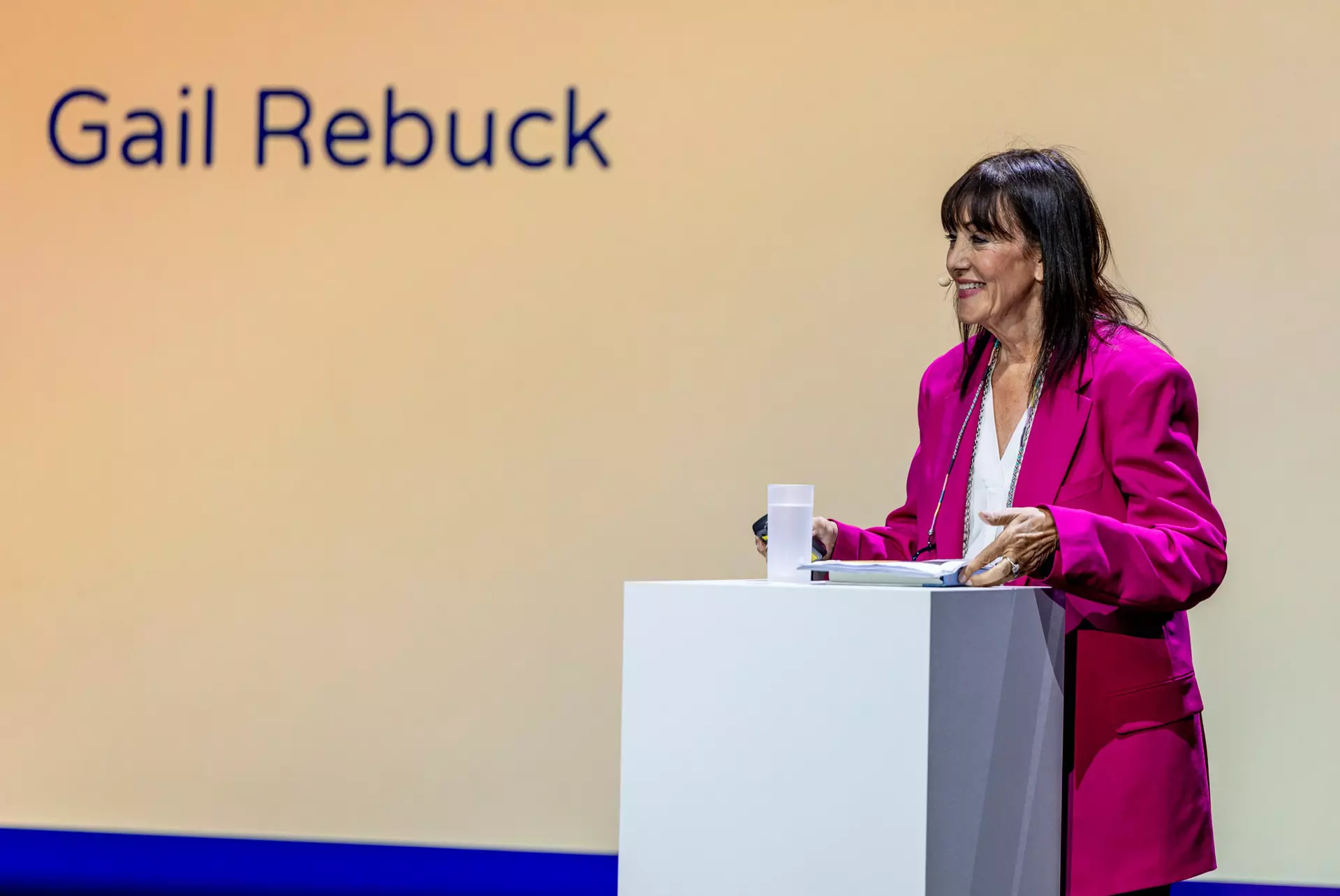 Person im pinken Anzug steht am Rednerpult vor Leinwand mit Schriftzug „Gail Rebuck“.