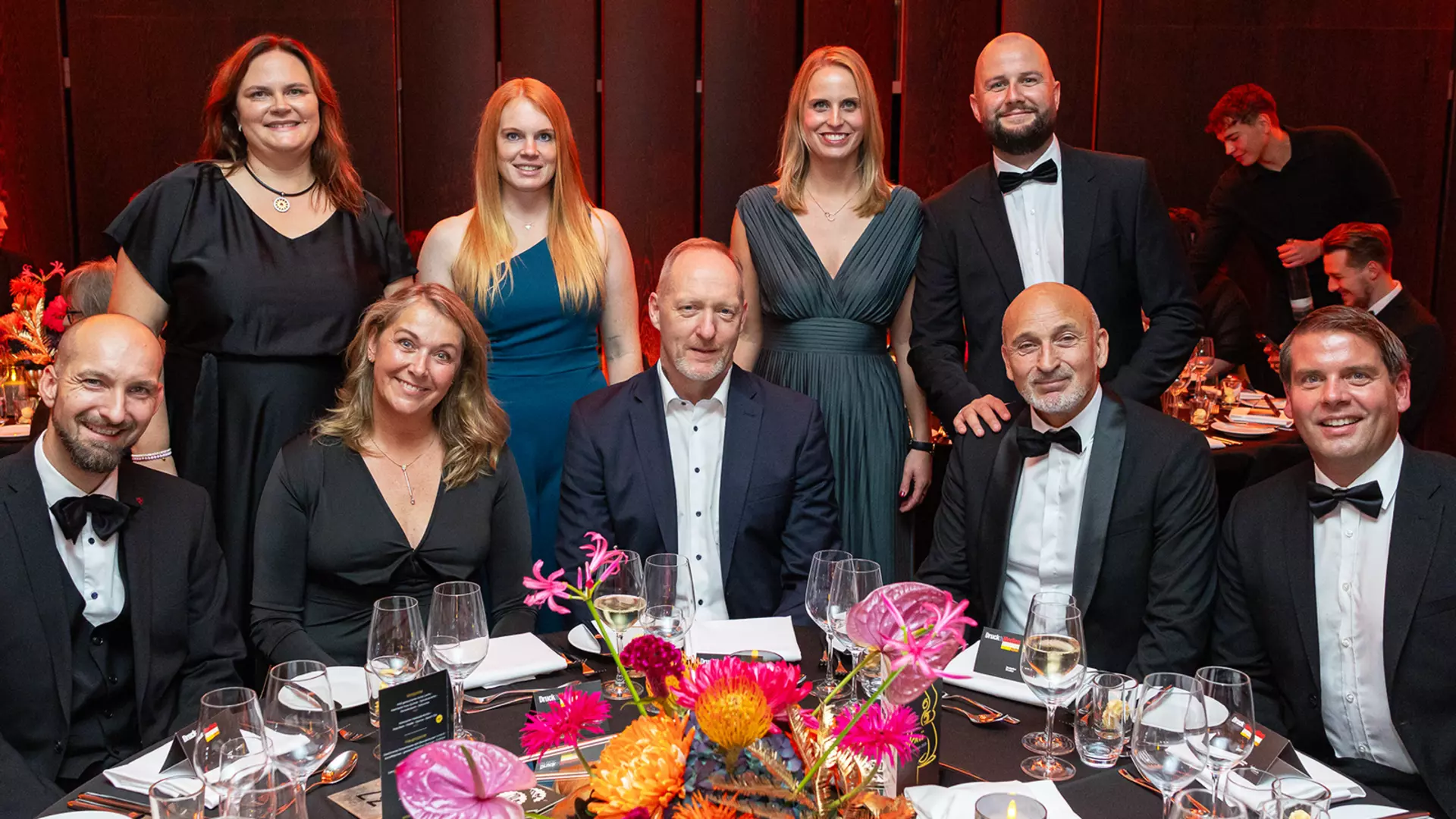 Group of people in formal evening attire sitting and standing around a festively set table with floral arrangements at a gala or dinner event.