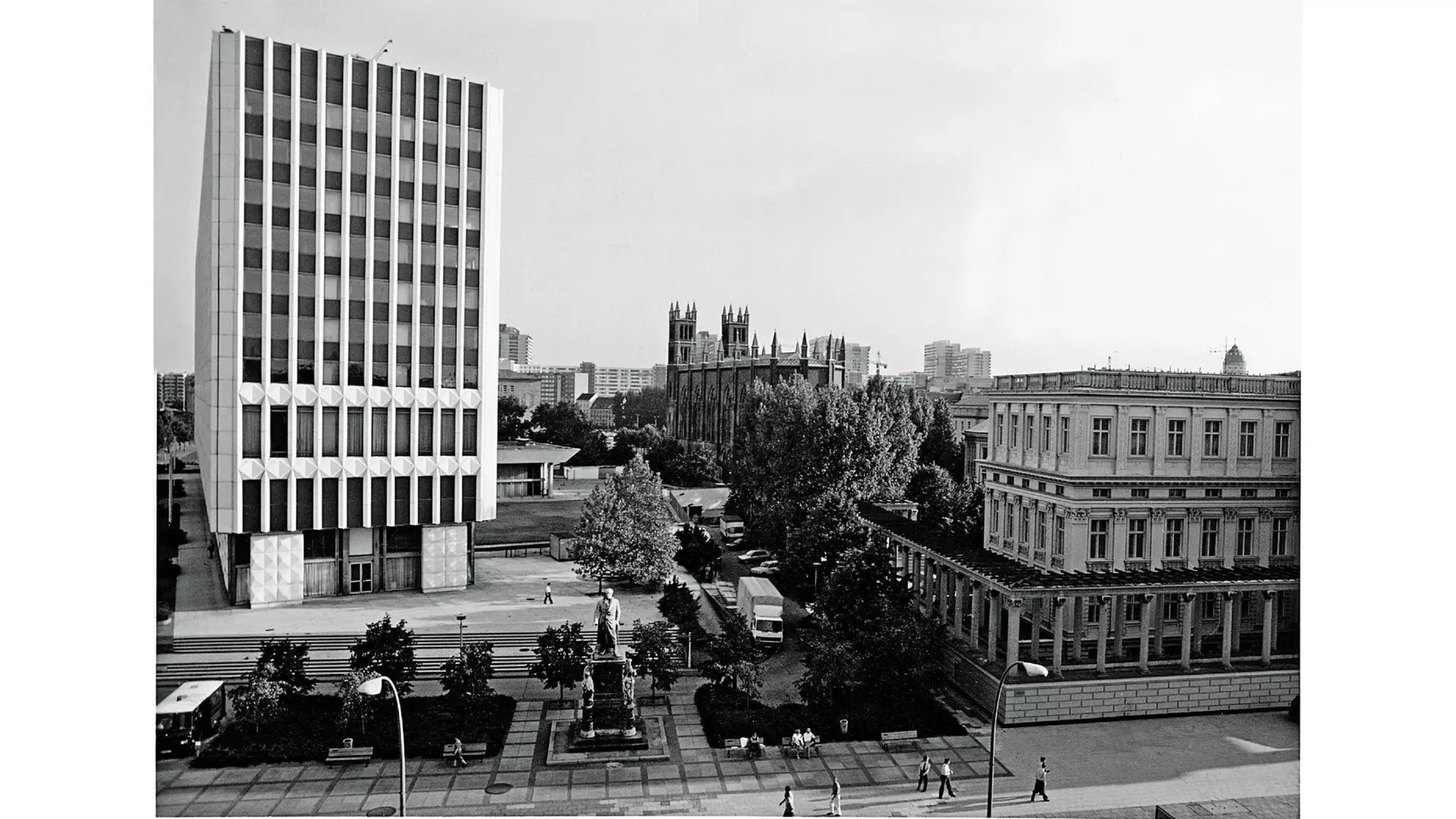 Historische Fotografie von 1972 mit modernem Hochhaus links, klassizistischem Gebäude rechts, dazwischen ein Platz mit Statue, Bäumen und Passanten.