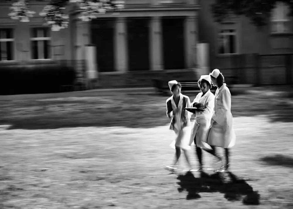 Drei Pflegekräfte in Uniform gehen über Rasen vor Gebäude mit Säulenfassade.