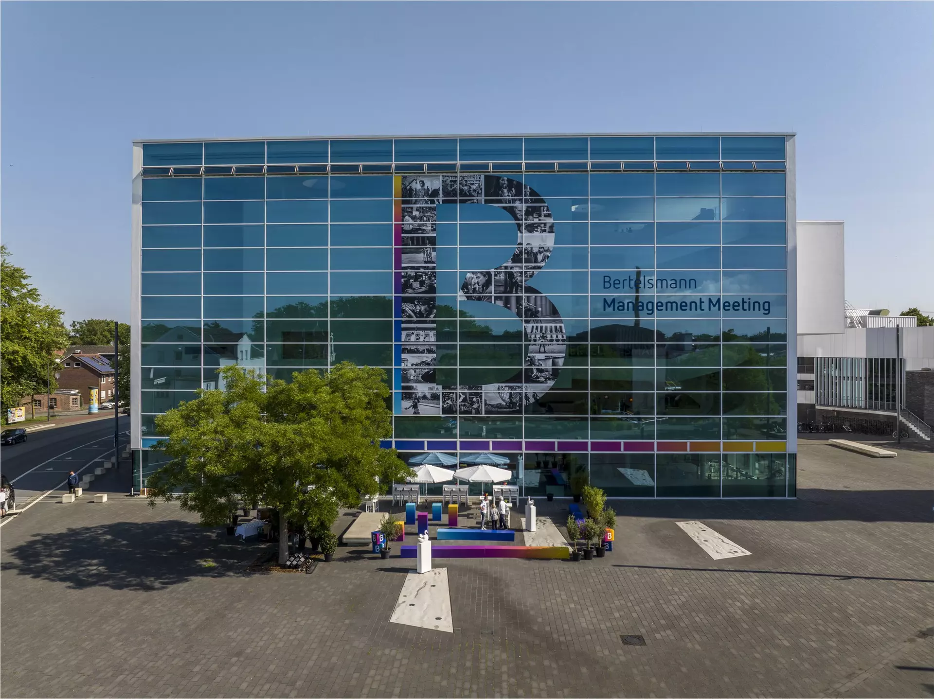 Glasfassade mit großem „B“ und Schriftzug „Bertelsmann Management Meeting“ vor modernem Gebäude.