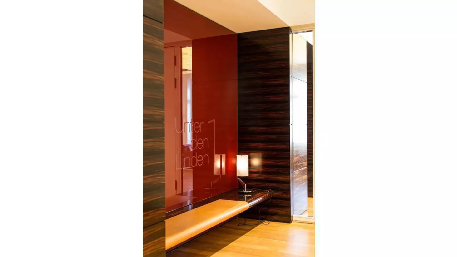 Moderner Innenraum mit Sitzbank, einer Leuchte und dunkelroter Glaswand; Schriftzug „Unter den Linden 1“ sichtbar, heller Holzfußboden und dunkle Wandpaneele.