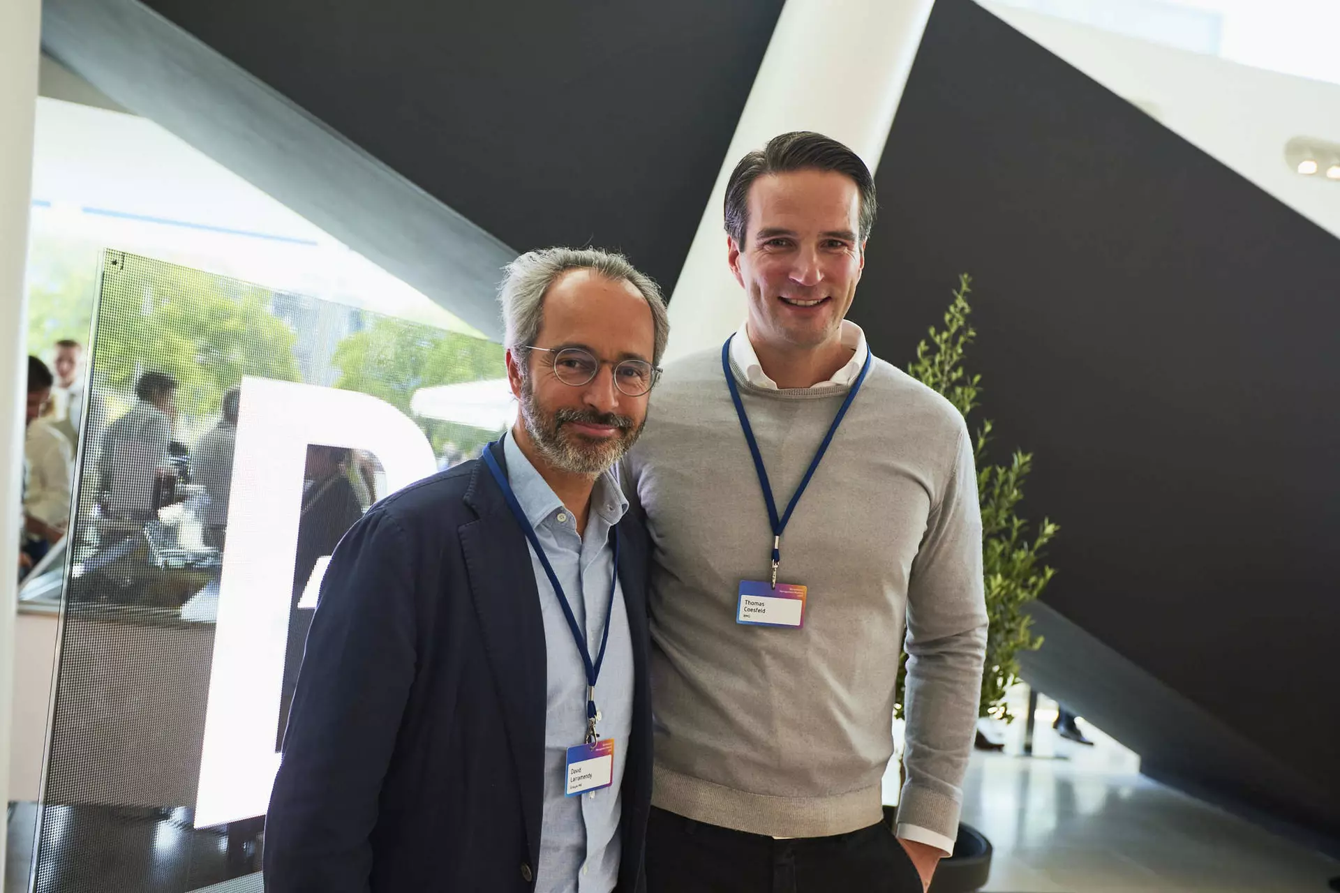 Zwei Personen mit Namensschildern stehen nebeneinander in modernem Foyer mit großem Buchstaben „B“ im Hintergrund.