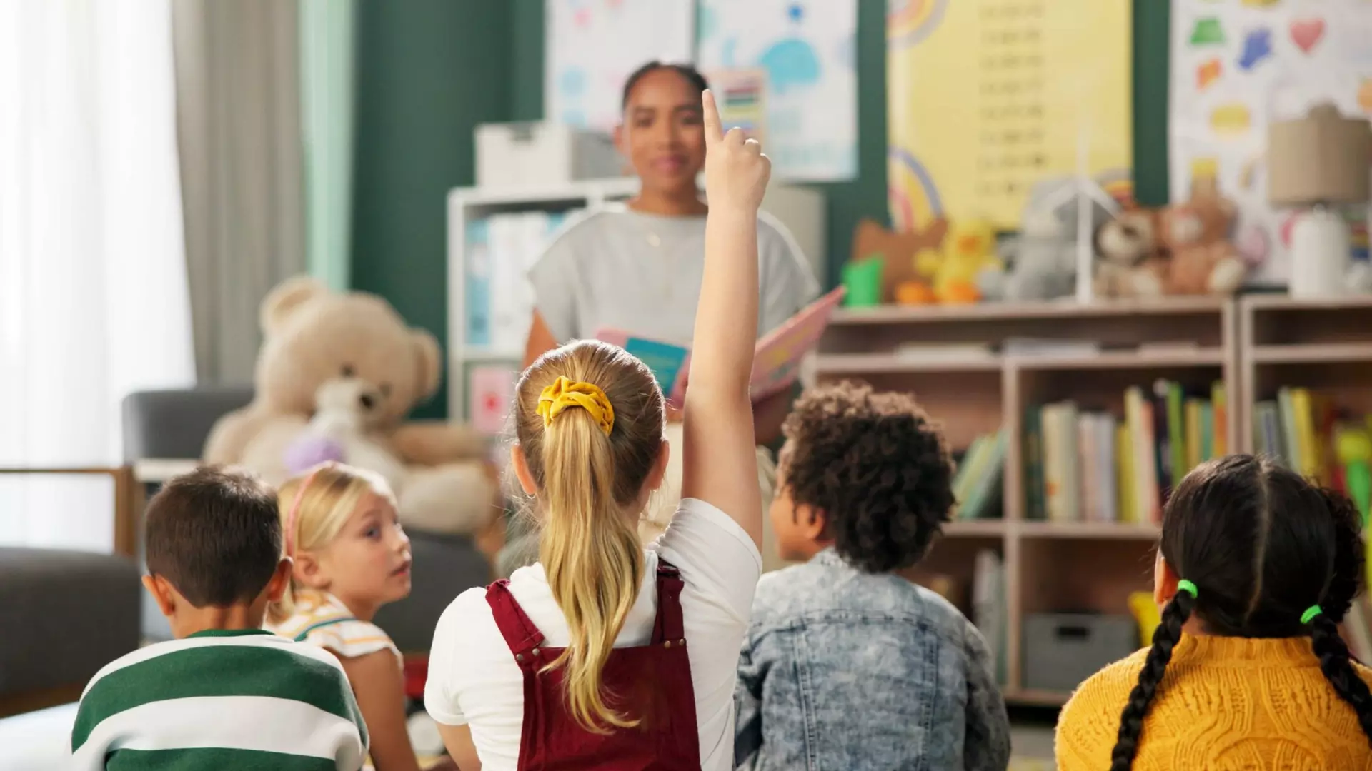 Children seated in a classroom facing a person reading aloud; one child raises a hand, with shelves and learning materials in the background.