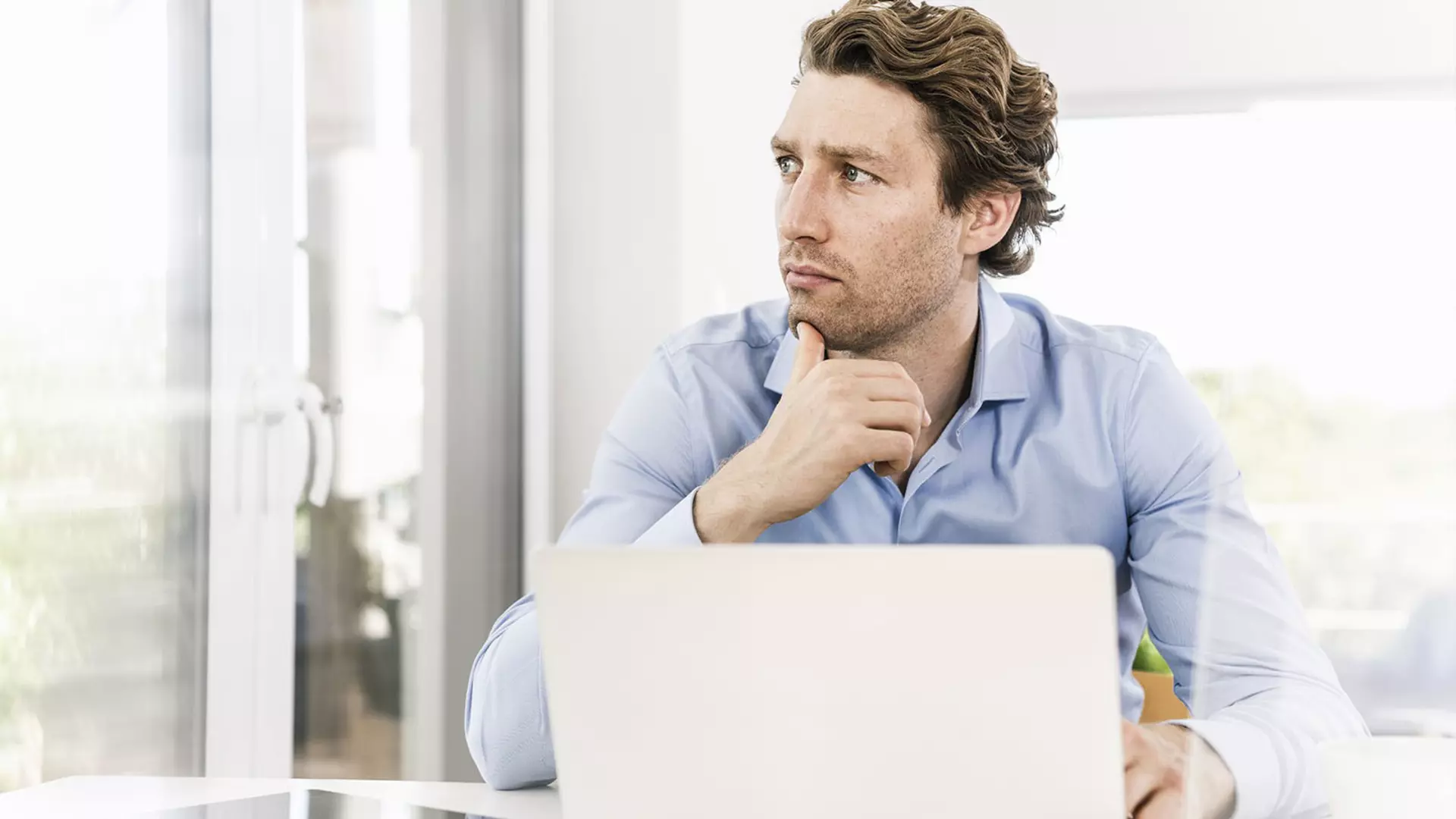 A man is looking thoughtfully to the side, sitting in front of an open laptop