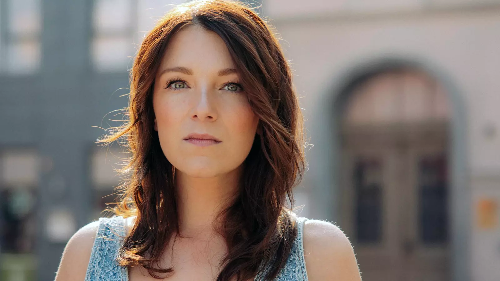 Person wearing a sleeveless top stands outdoors in front of a blurred building, captured in natural light.