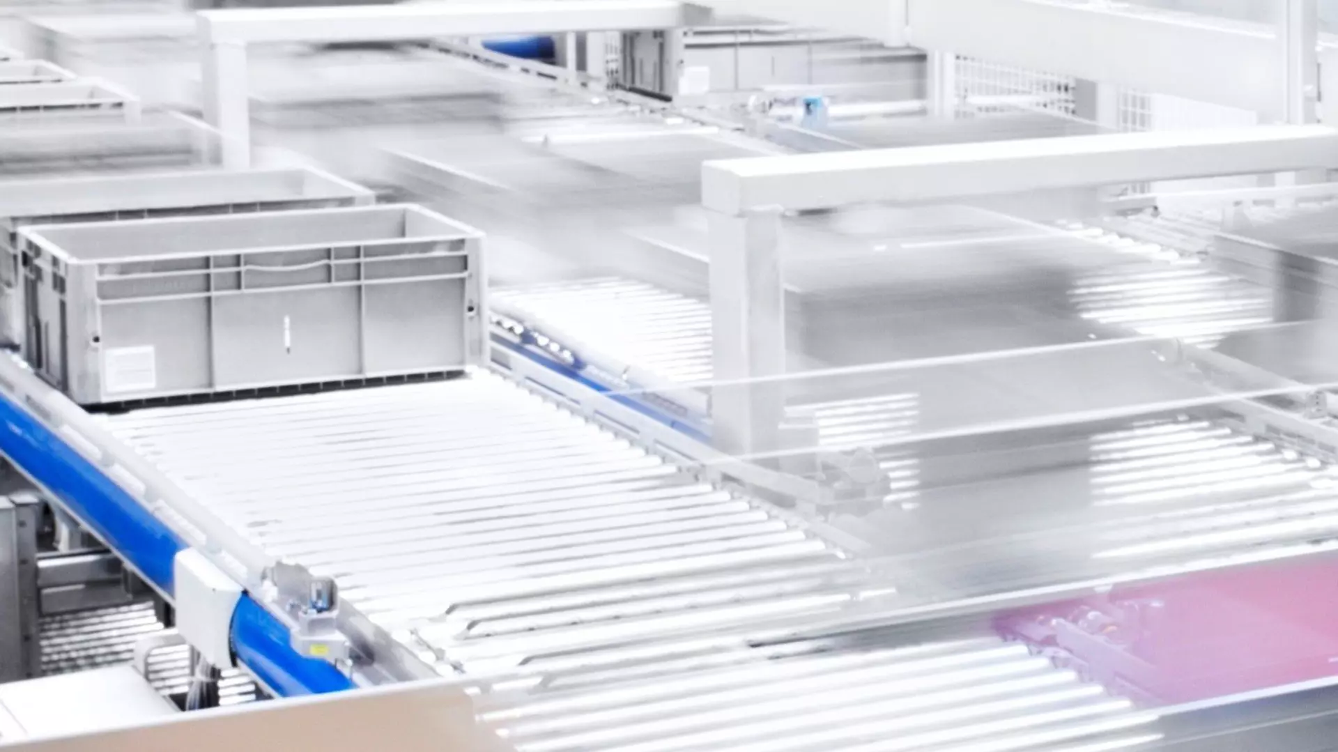 An automated conveyor belt system transports gray plastic containers through a modern logistics hall.