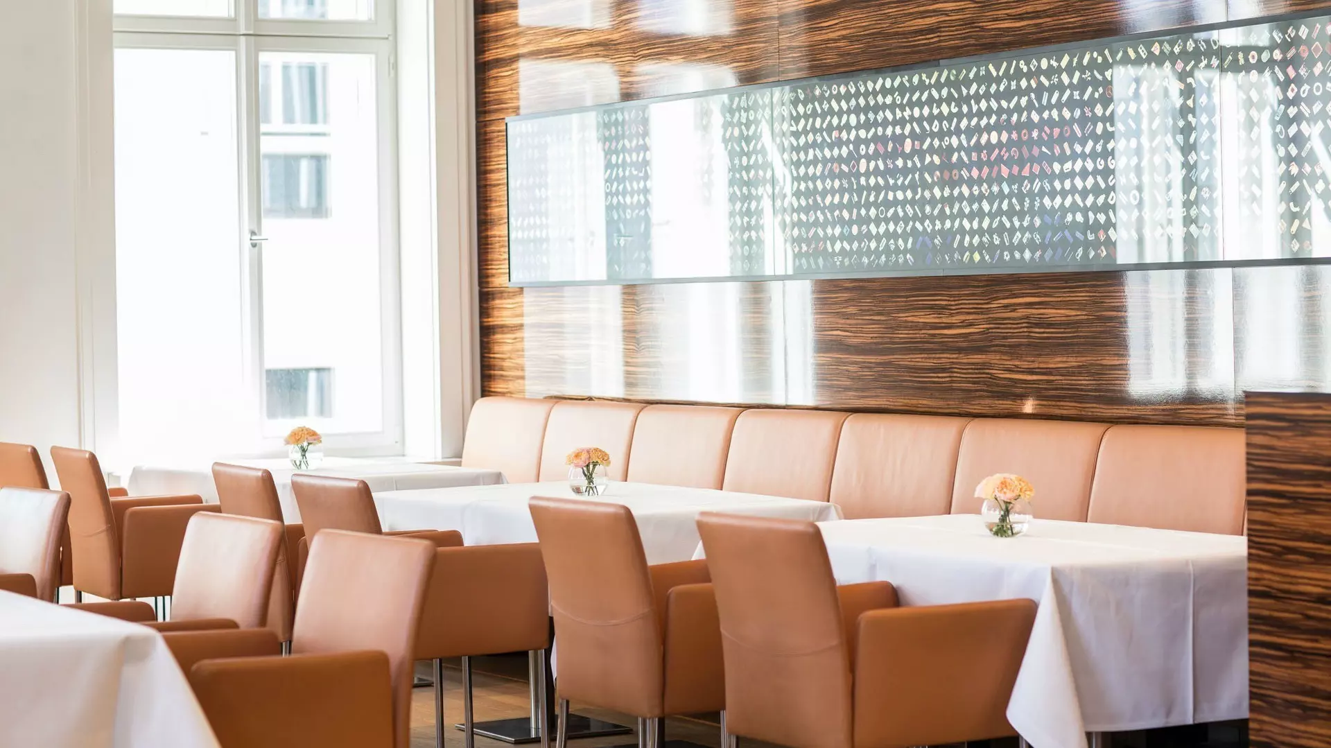 Restaurantbereich mit gedeckten Tischen, lederbezogenen Stühlen, Sitzbank an Holzwand, daran ein langes, verglastes Bild und Tageslicht durch Fenster.