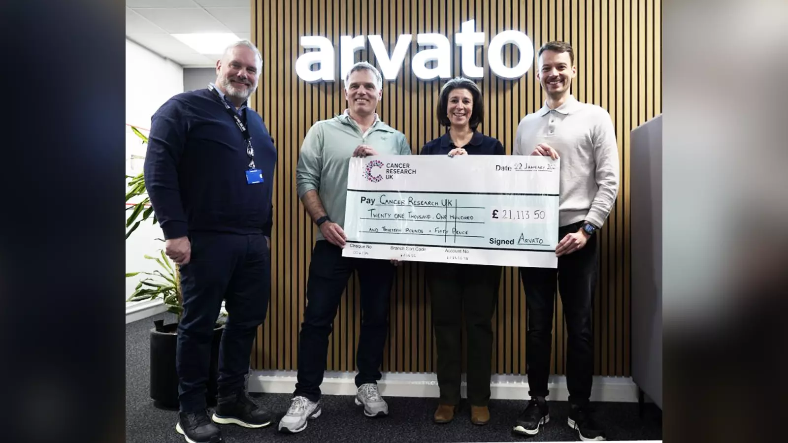 Four people standing in front of a wooden slat wall with the Arvato logo, holding a large donation cheque for Cancer Research UK.