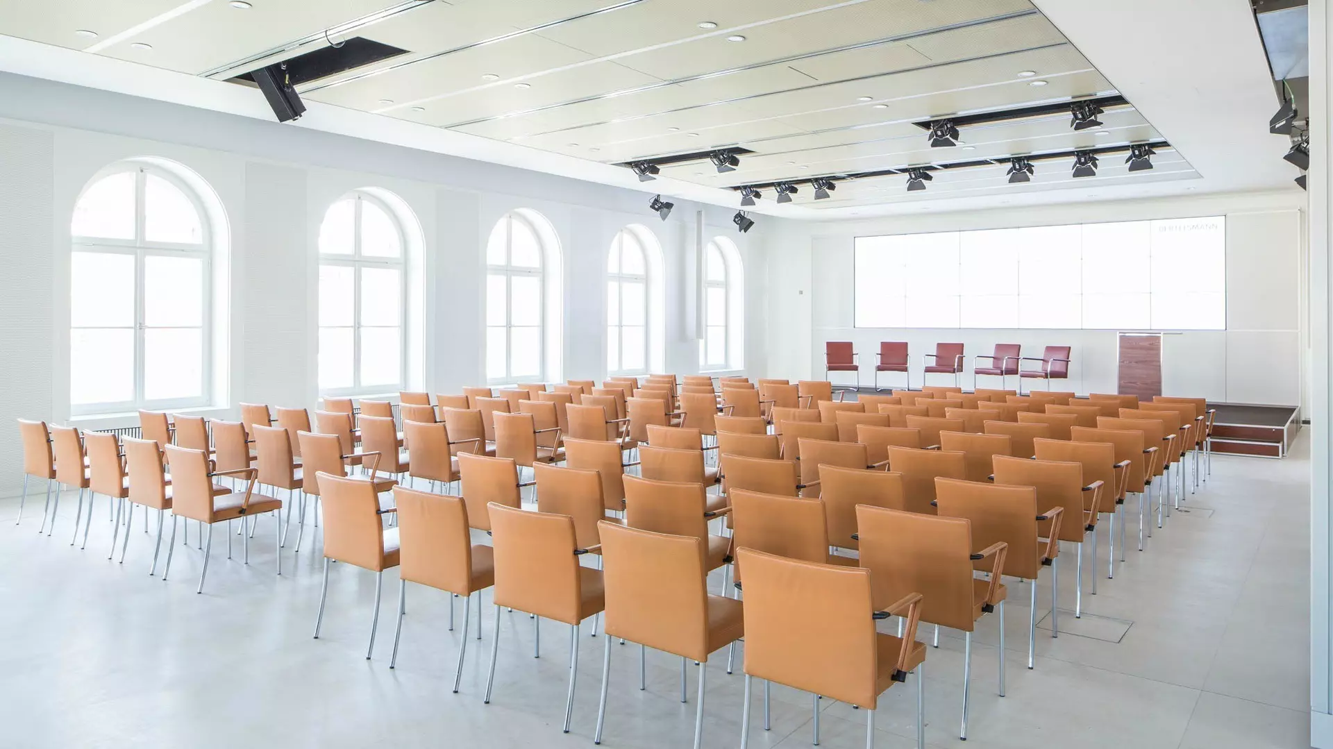 Großer Veranstaltungsraum mit Reihen hellbrauner Stühle, Bühne mit Podium und großer LED-Rückwand, hohen Fenstern und moderner Deckenbeleuchtung.