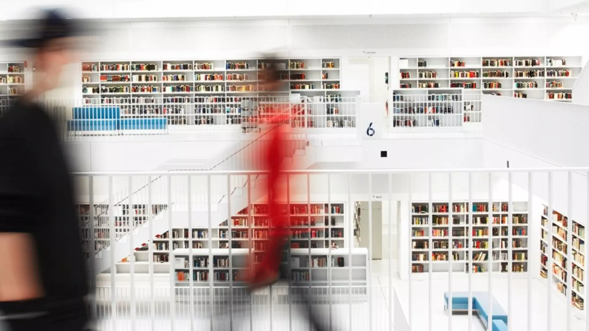 In the foreground, two blurred figures in black and red can be seen, with a large library with steps in the background.