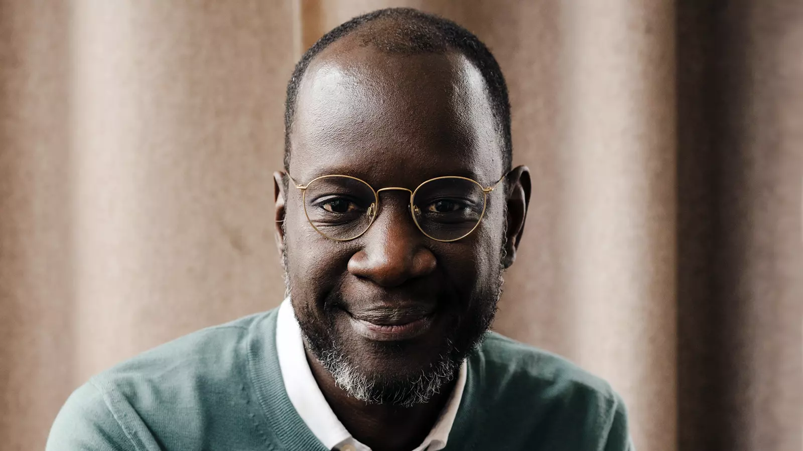 Person wearing a light shirt and knit sweater sits indoors in front of curtains and looks forward.