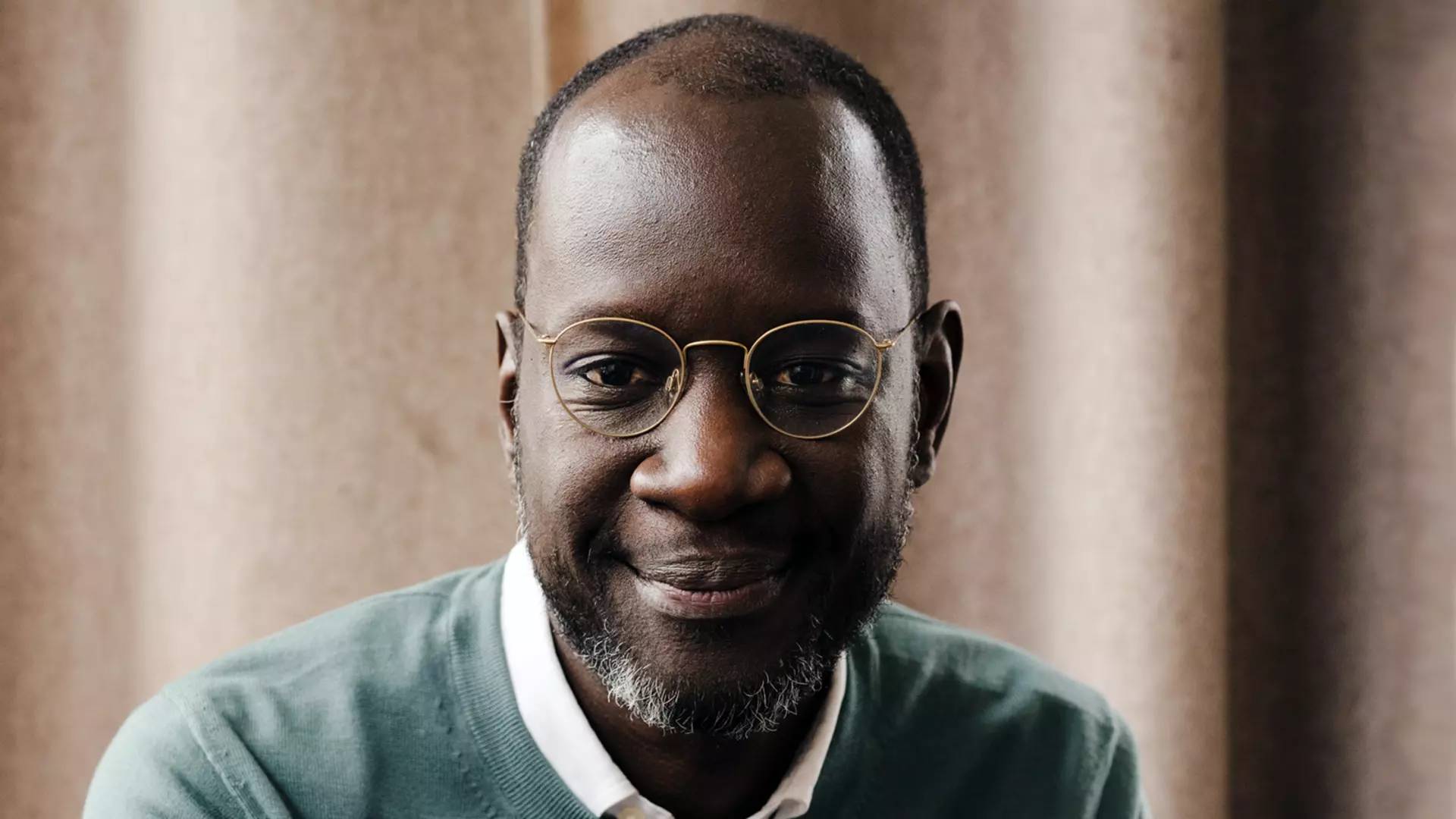 Person wearing a light shirt and knit sweater sits indoors in front of curtains and looks forward.