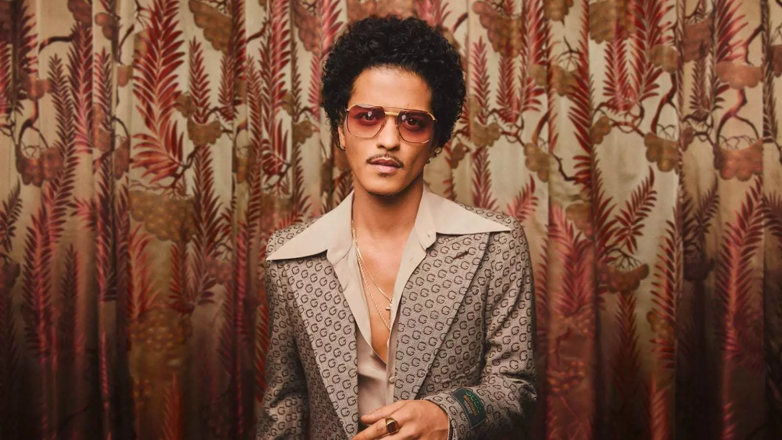 The singer Bruno Mars stands in a suit in front of a patterned curtain