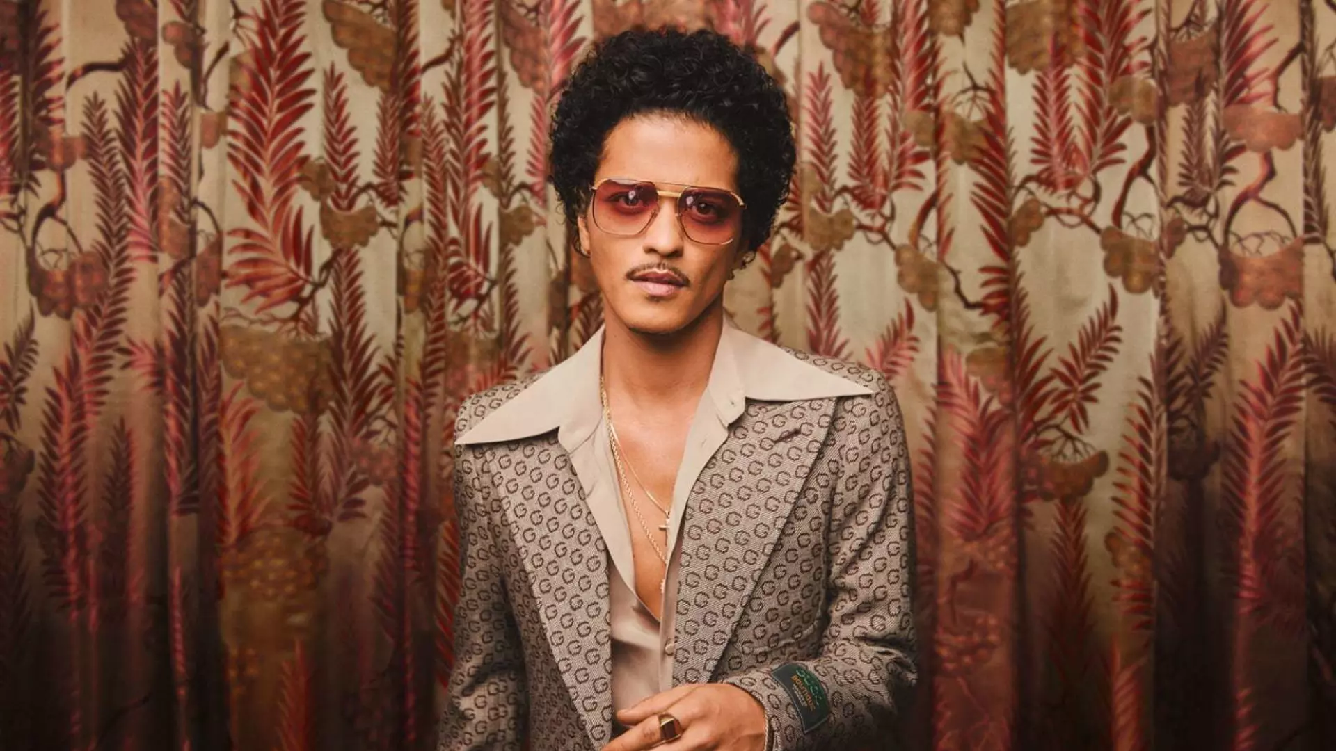 The singer Bruno Mars stands in a suit in front of a patterned curtain