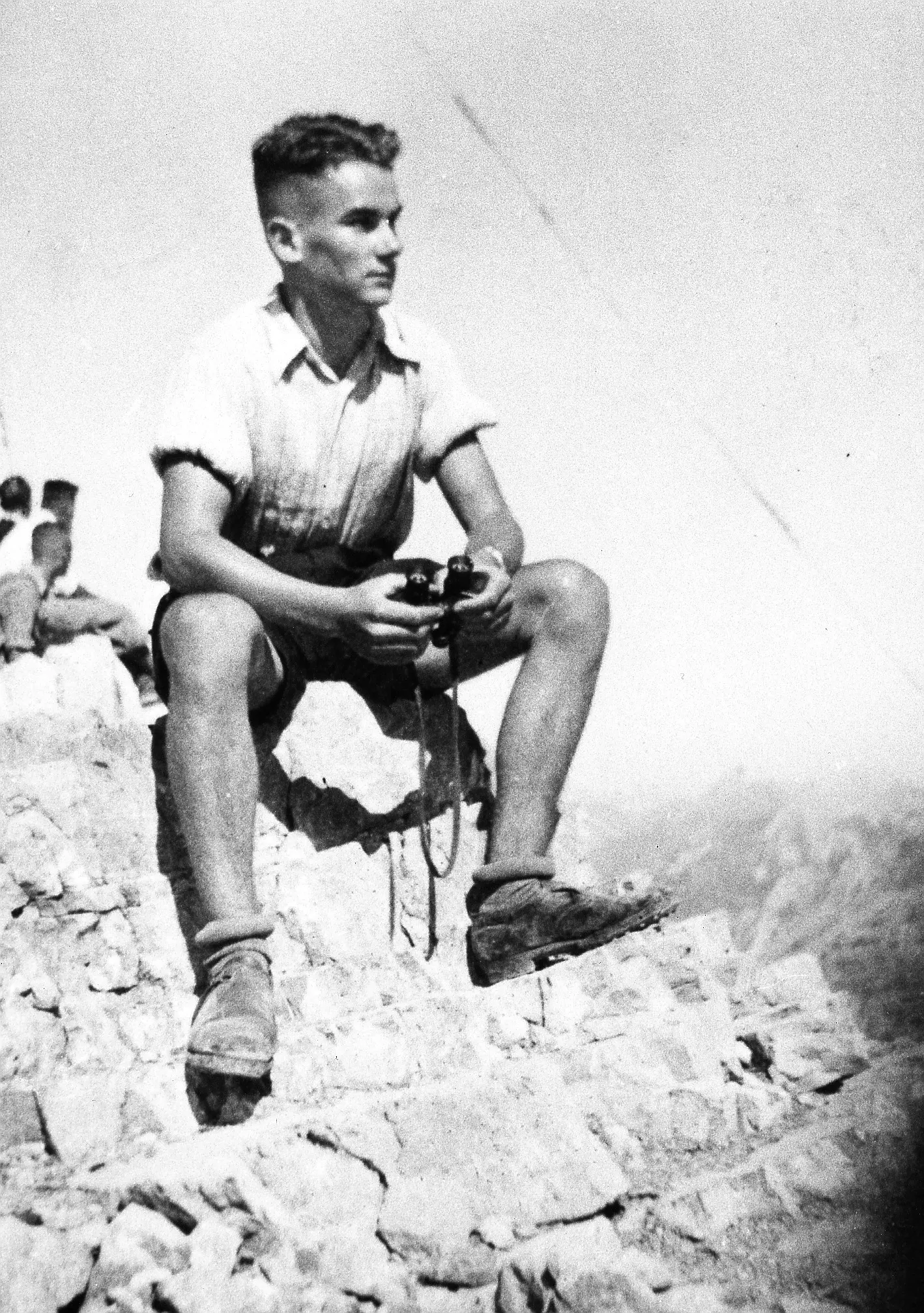 The young Reinhard Mohn sits on a rock in the mountains with binoculars in his hand.